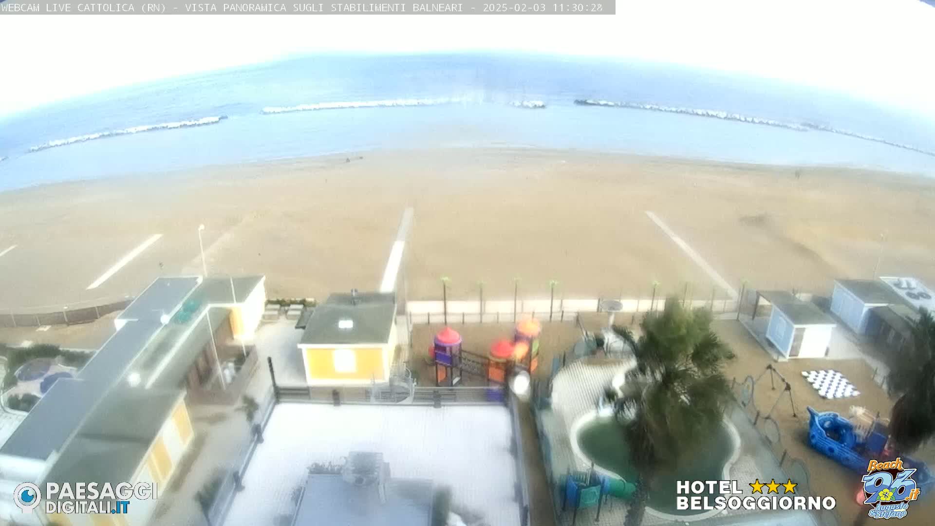 An overcast day shows a sandy beach with calm waves, beach structures, and a playground near the water.