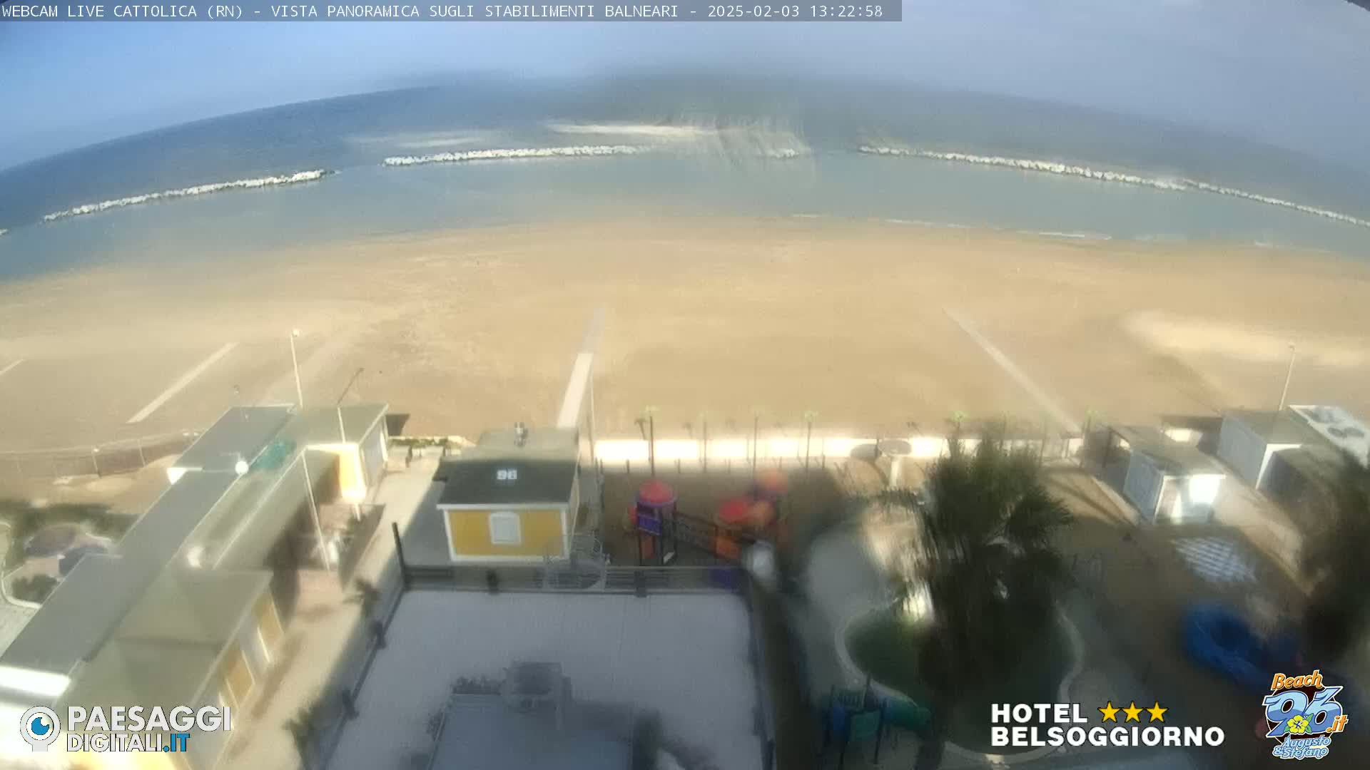 An aerial view shows a sandy beach, calm ocean, and beachfront buildings, including a playground, on a partly sunny day.
