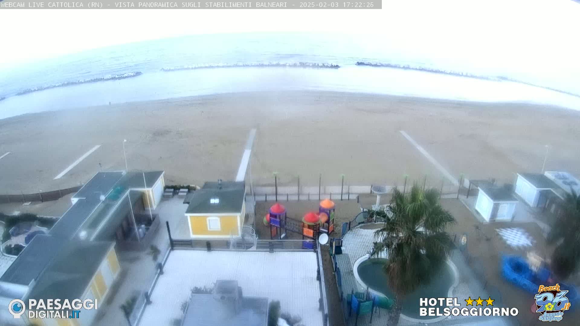 An overcast day shows a beach with light waves, and a view of beach buildings including a playground and swimming pool from an elevated perspective.