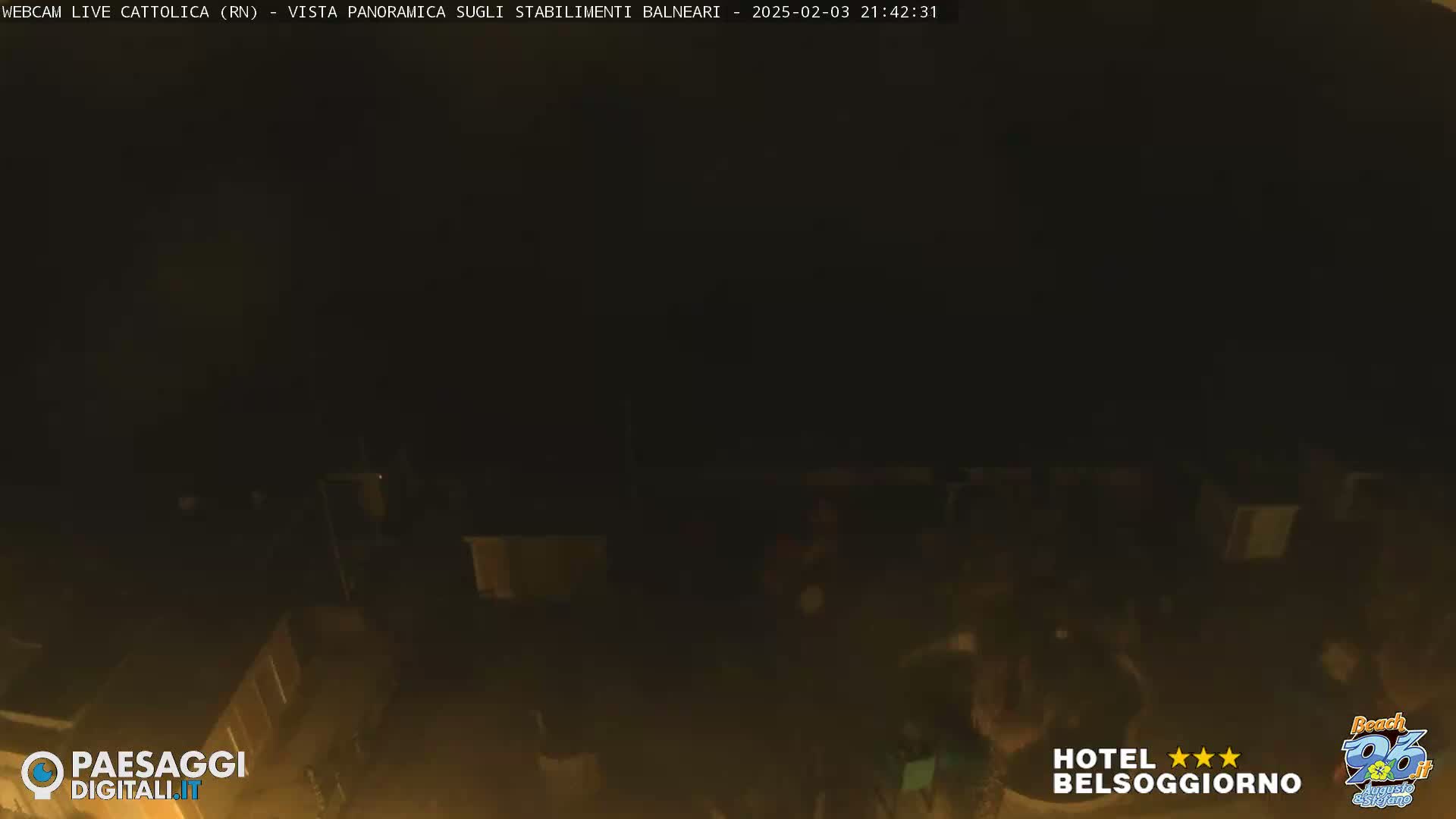 A nighttime, dark view of a beach area with dimly lit buildings and some indistinct figures.