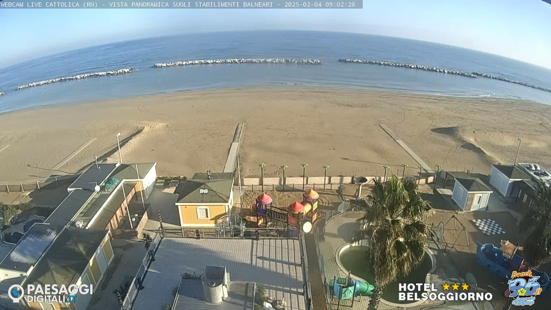 An aerial view shows a sandy beach with calm ocean water under a clear sky, beachside buildings, a playground, and a small pool.
