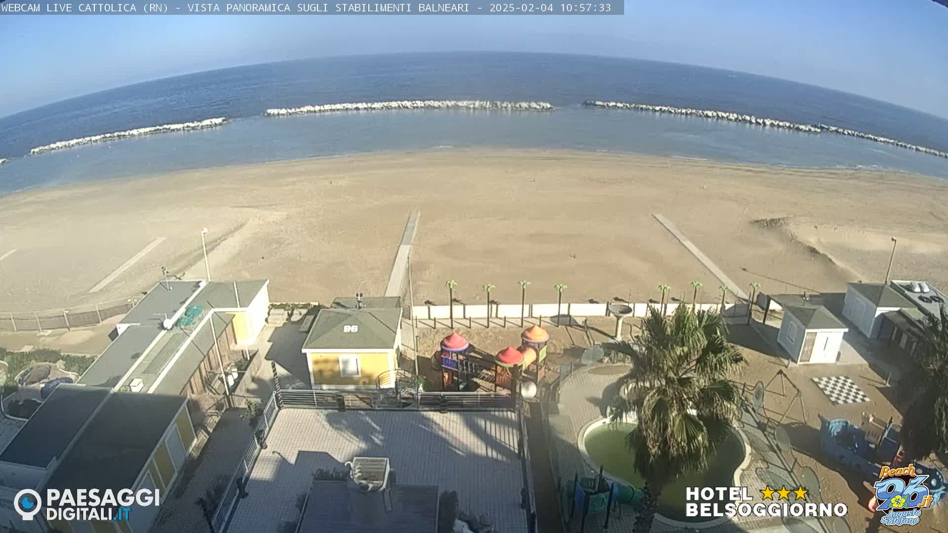 A sandy beach with calm ocean water and a row of breakwaters, viewed from an elevated position showing beachside buildings, a playground, and a pool.