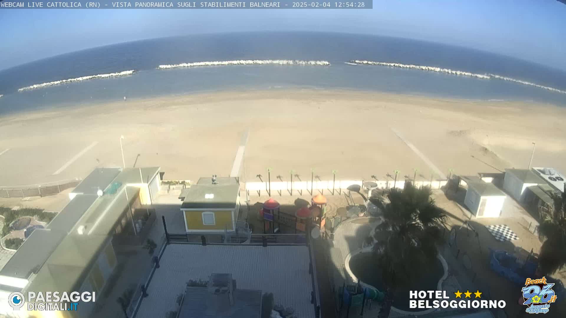 A panoramic view of a sandy beach, calm sea with breakwaters, and beachside buildings including a playground, under sunny skies.