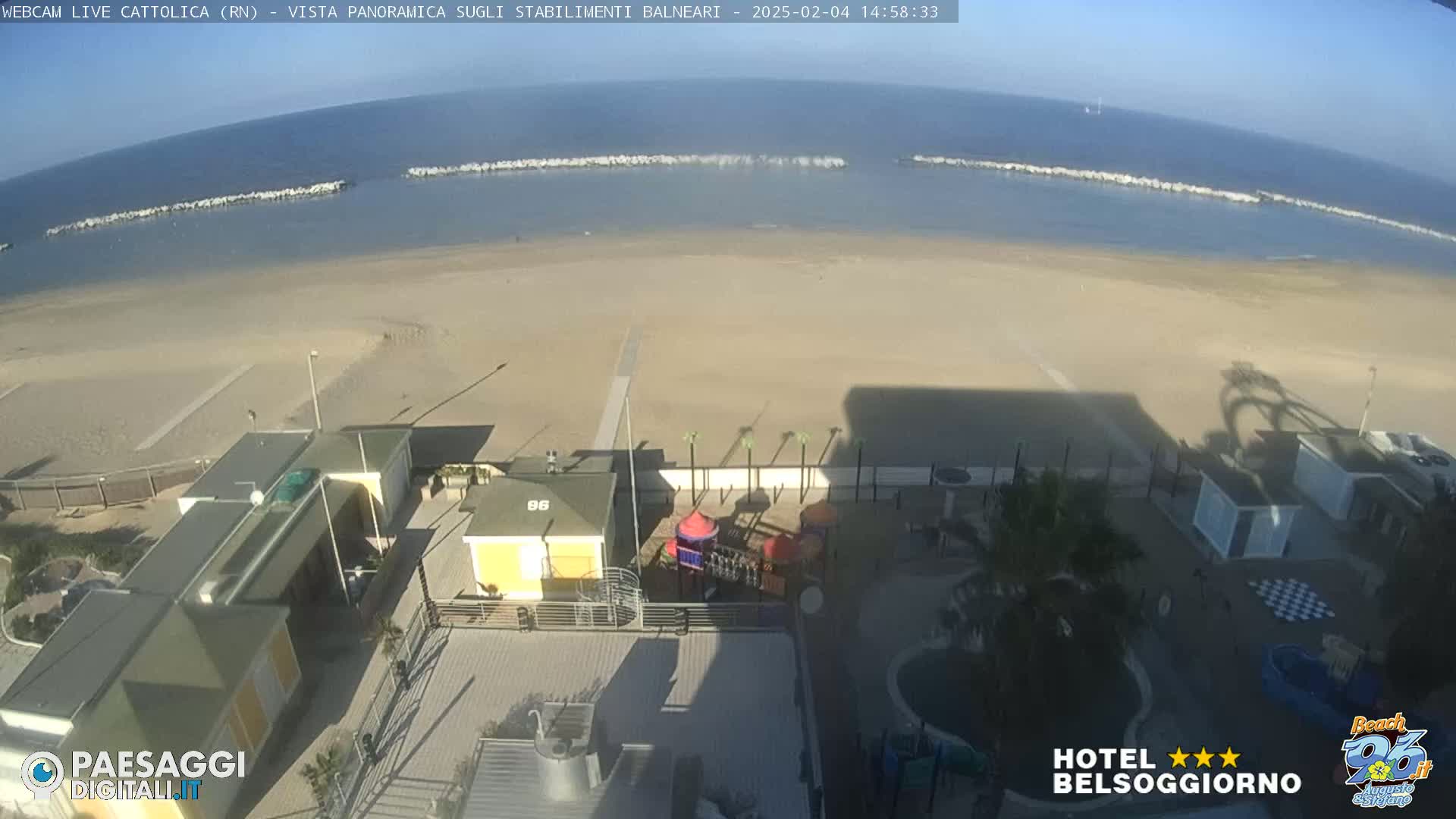 A high-angle, sunny view of a beach with calm water, breakwaters, beachside buildings, and a playground.