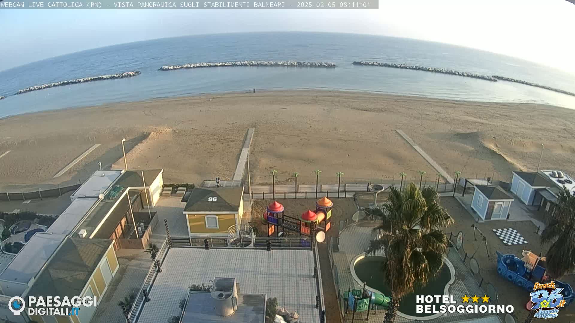 A beach with calm water, breakwaters, and beachside buildings including a playground and pool, under a clear sky.
