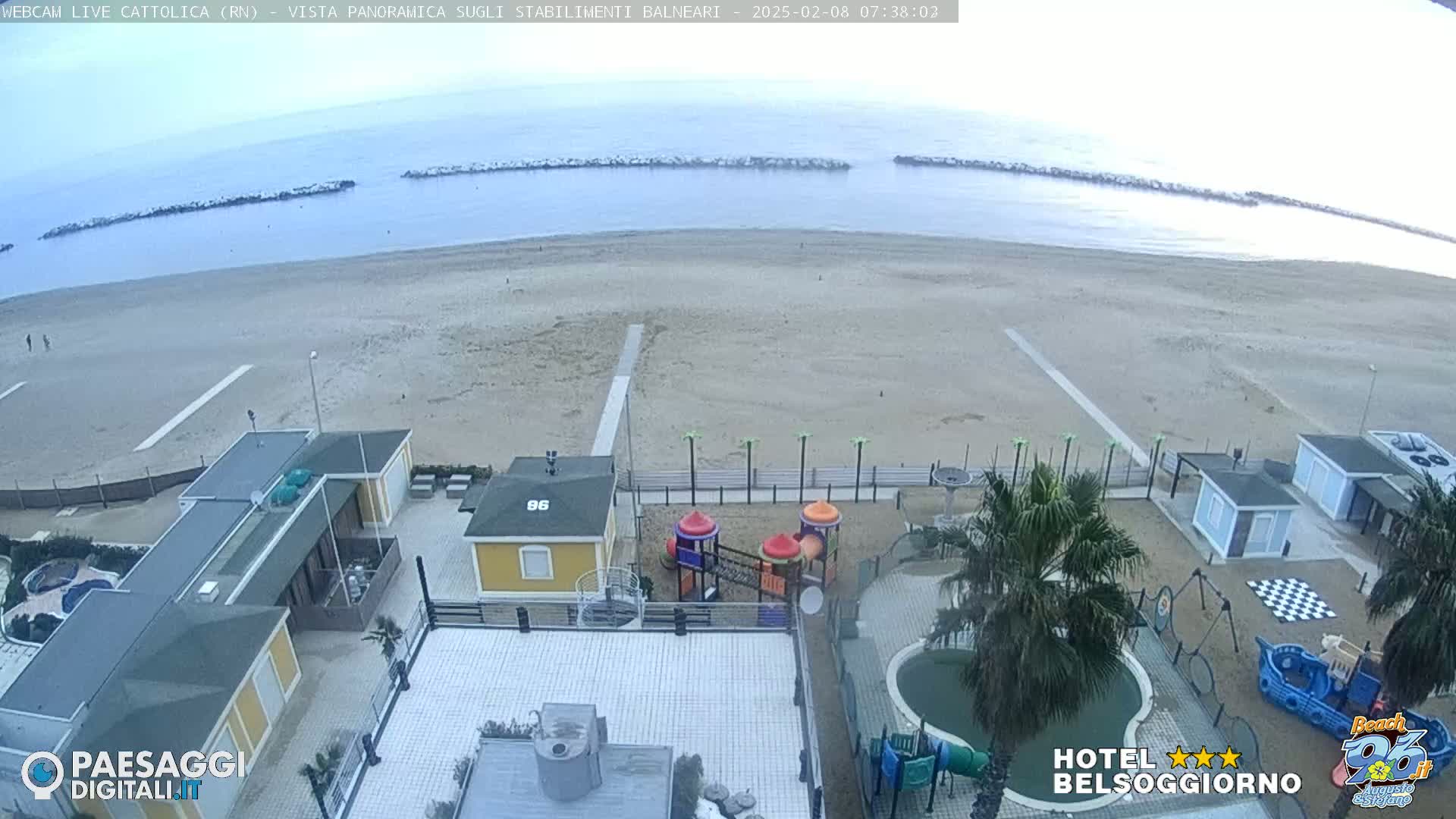 An overcast view of a sandy beach with calm water, beachside buildings, a playground, and a small pool.