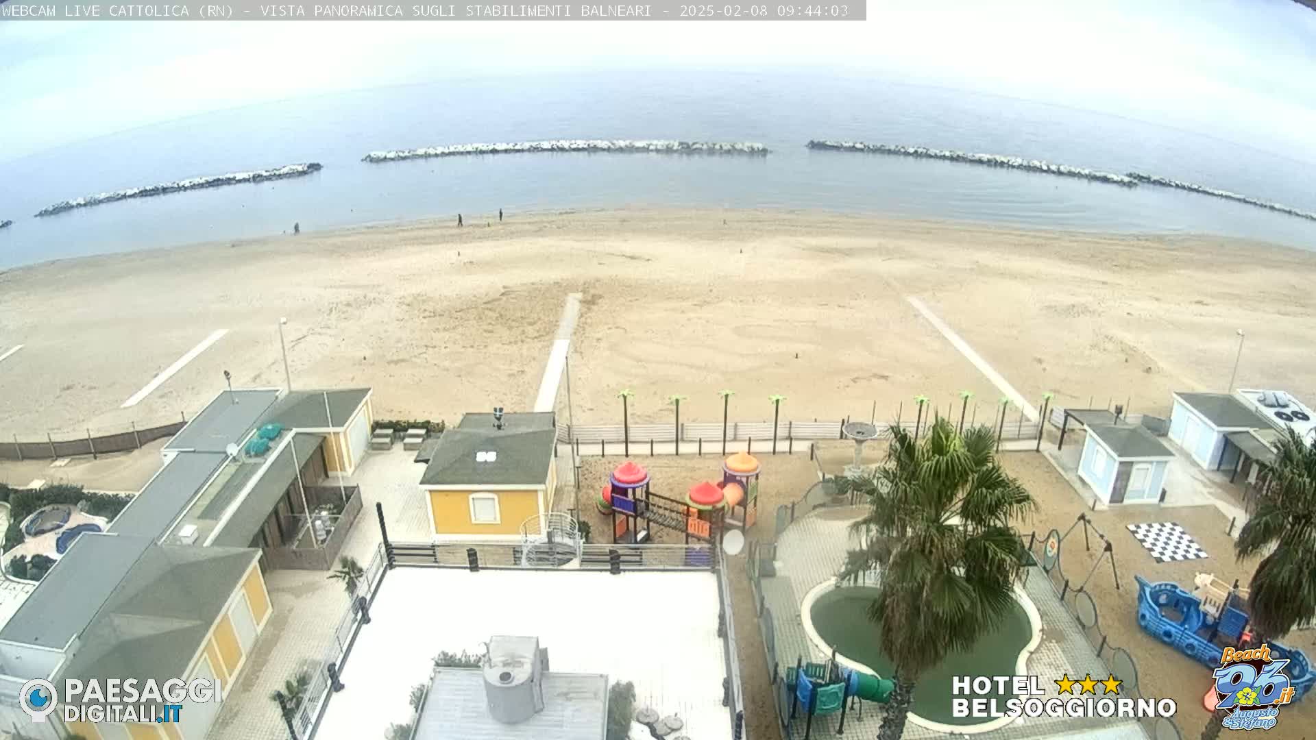 An overhead view of a sandy beach with calm ocean water, beachside buildings, a playground, and a small pool under an overcast sky.