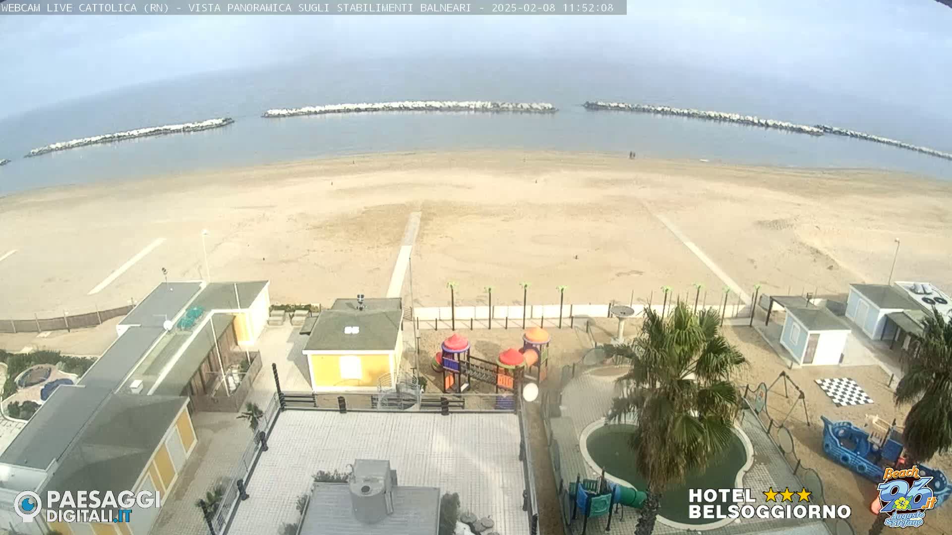 An overhead view shows a sandy beach with calm water and breakwaters, overlooking a beachfront area with buildings, a playground, and a small pool under a hazy sky.
