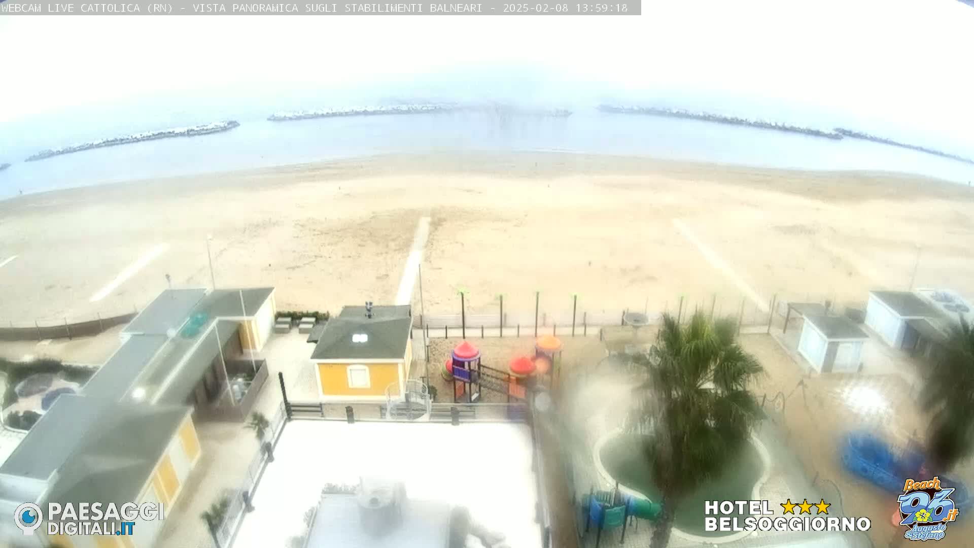 A beach with low-rise buildings, a playground, and a pool is visible from an elevated vantage point on an overcast day.