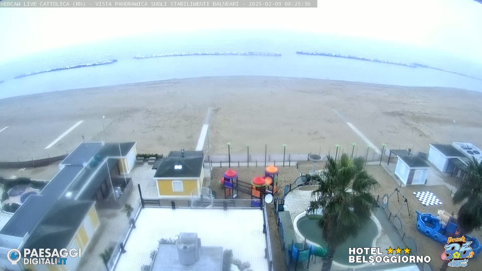 A mostly cloudy day at a beach shows a sandy shoreline, calm ocean water, beachfront buildings, a playground, and a swimming pool.
