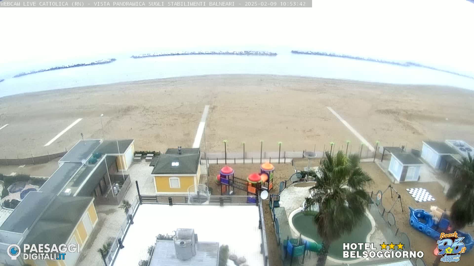 A beach with calm, overcast water is viewed from above, showing beachside buildings, a playground, and a small pool.