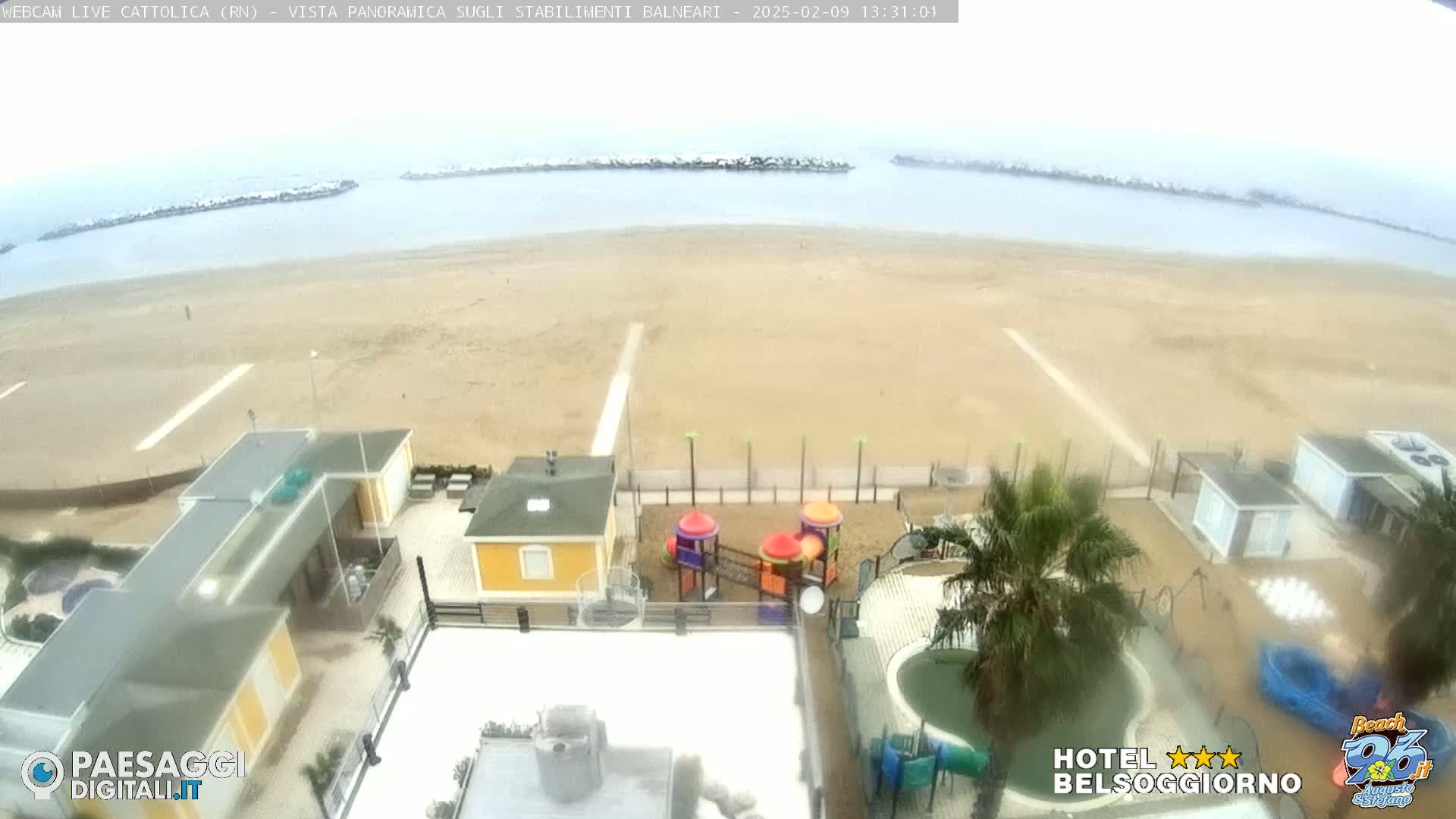 A beach with overcast skies is viewed from above, showing beach structures, a playground, and a pool.