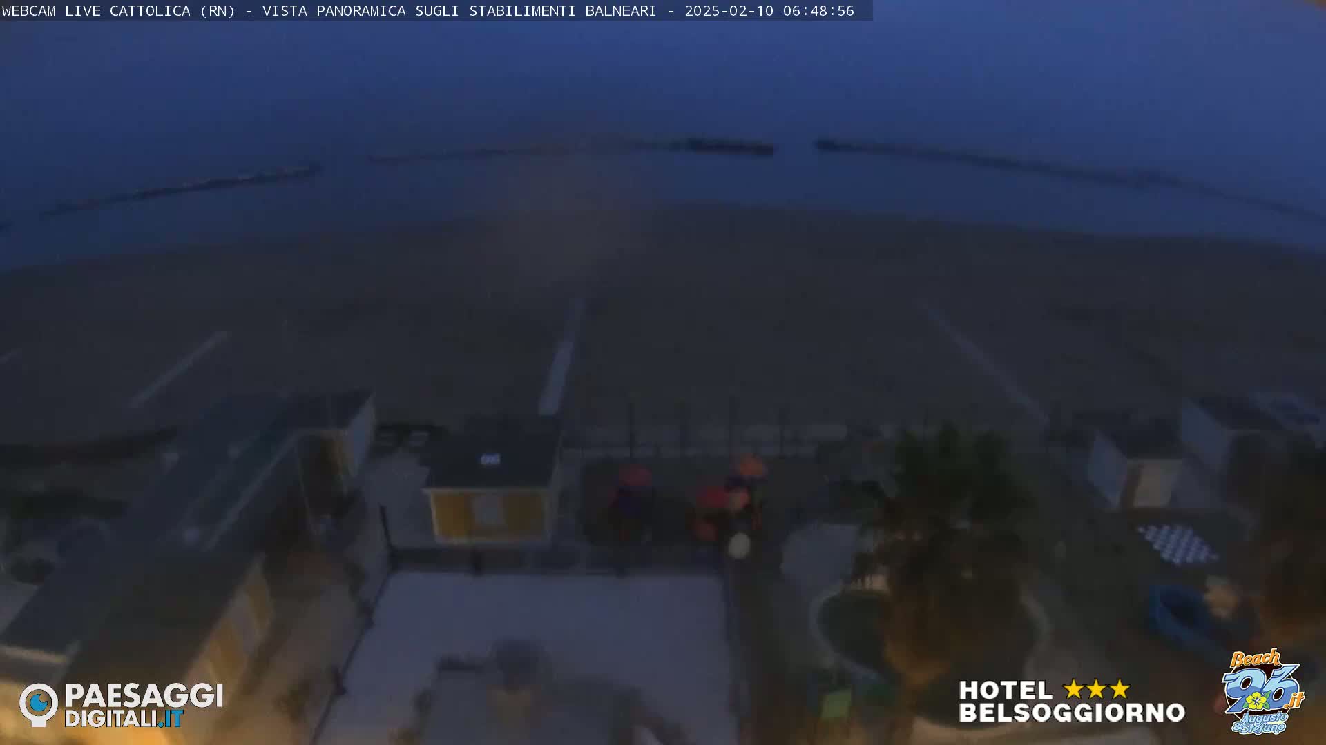 A dark, pre-dawn view shows a beach, calm sea, and a collection of low buildings near the water.