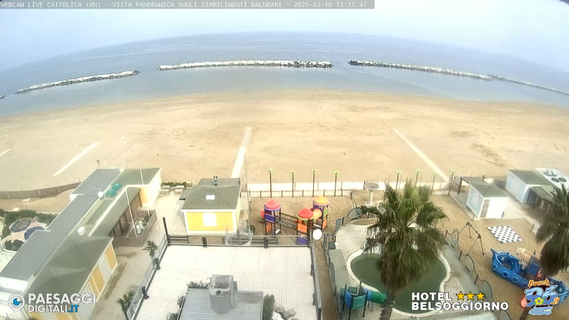 A sandy beach stretches to a calm sea under an overcast sky, with beachside buildings and a playground visible.