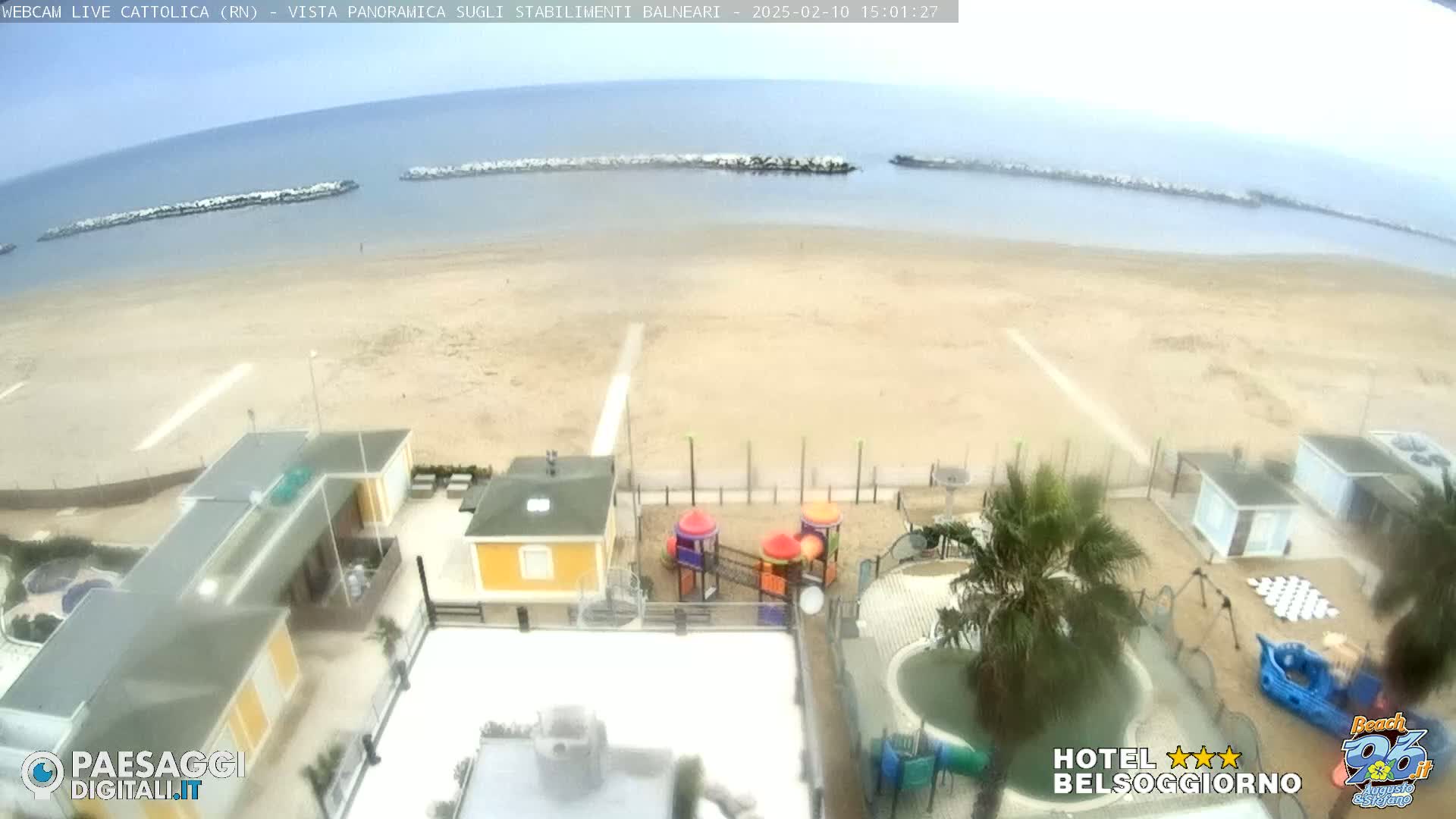 An overcast day at a beach shows a sandy shore, calm sea with breakwaters, beachside buildings, and a playground.