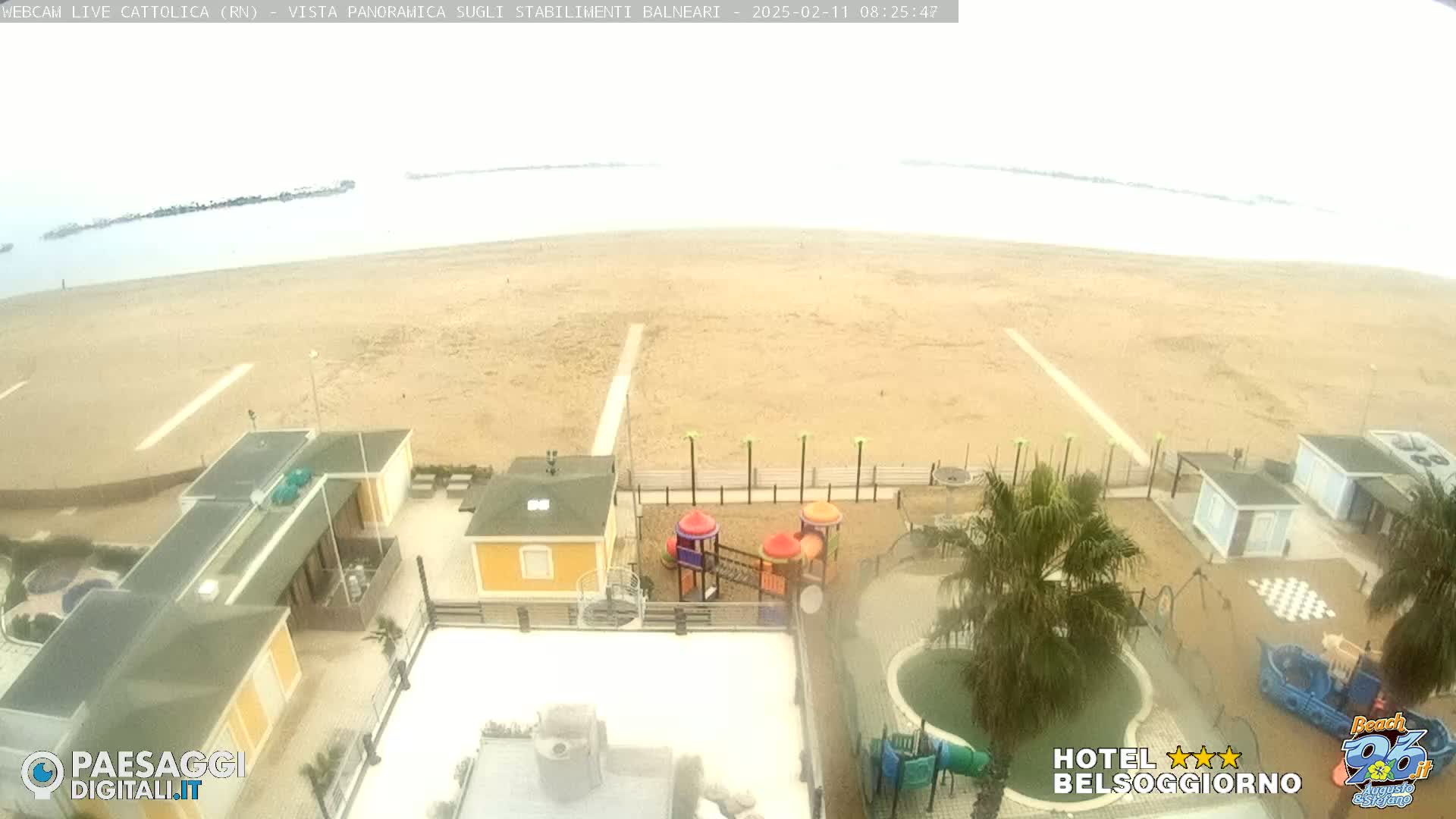 An overcast beach scene shows a sandy shoreline, calm water, beachside buildings, and a playground.
