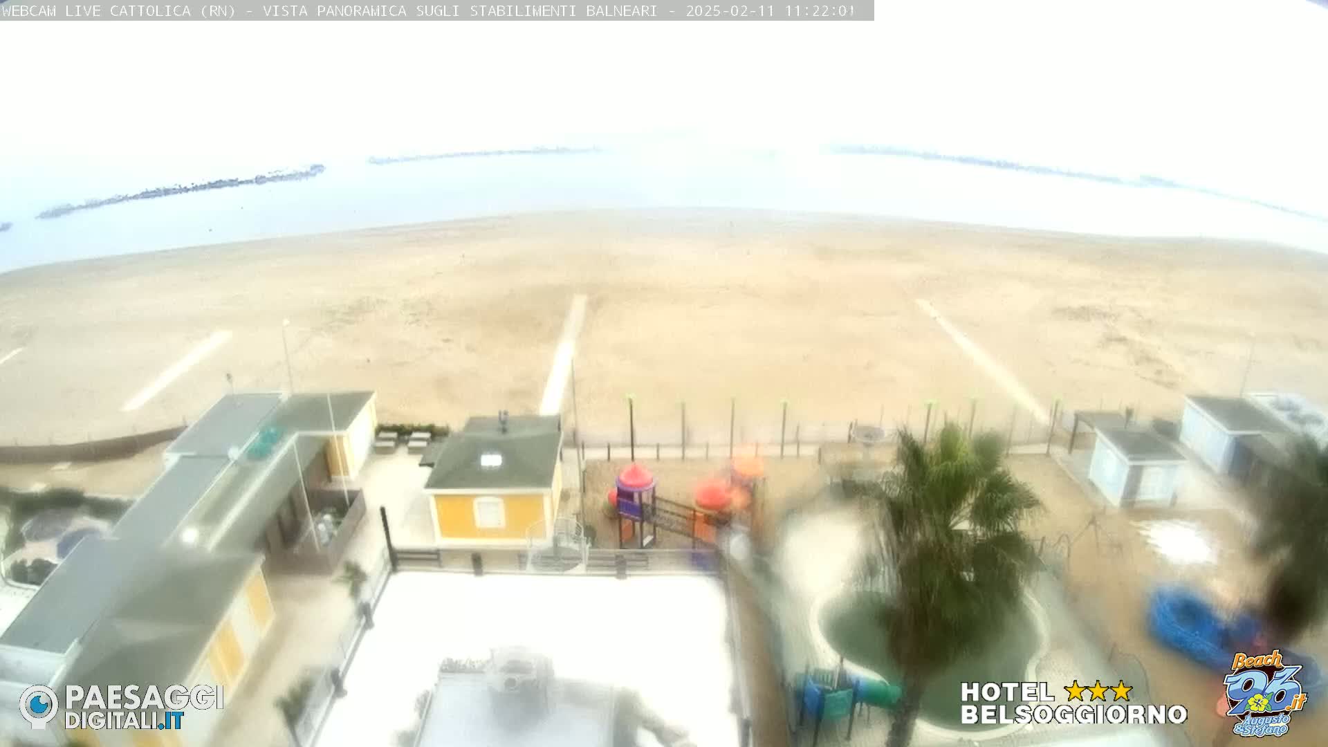 A beach with overcast skies is viewed from above, showing beach buildings, a playground, and a pool.
