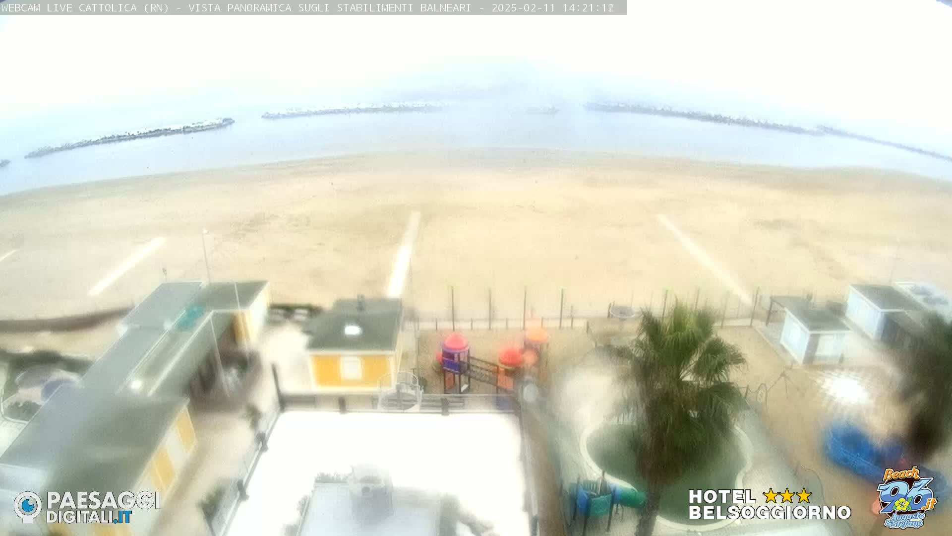 A hazy, overcast view of a sandy beach with beach houses, a playground, and a pool, overlooking calm ocean water with breakwaters.