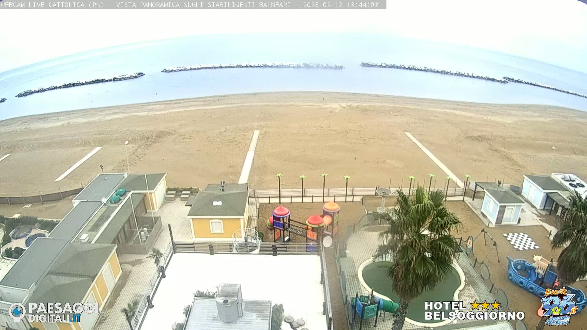 An overcast day at a beach shows a sandy shoreline, buildings, a playground, and a small pool, all near calm ocean water with breakwaters.