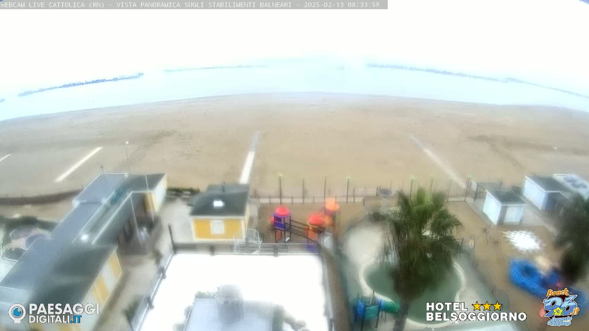 An overcast day at a beachside resort, showing a sandy beach, calm ocean, buildings, a playground, and a pool.