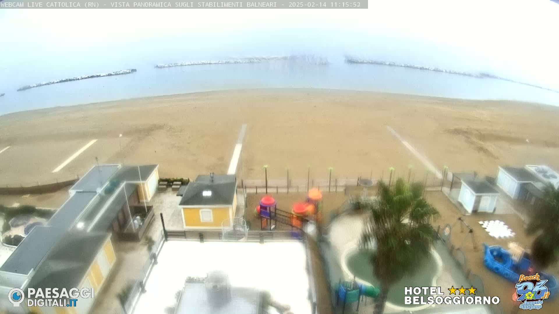 An overcast day at a beach shows a wide sandy shore with calm water and breakwaters, alongside beach buildings, a playground, and a small pool.