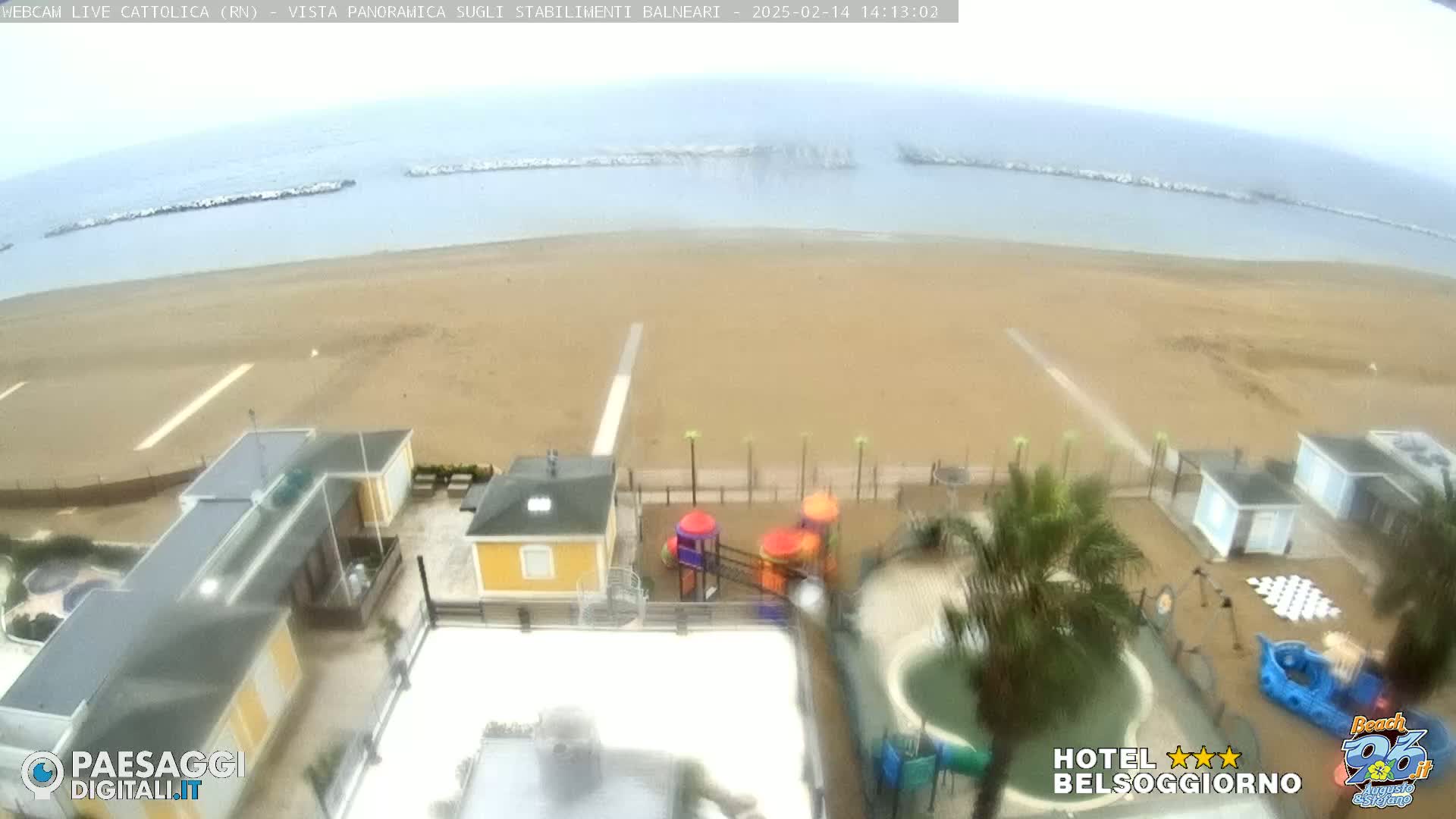 An overcast day shows a sandy beach with buildings, a playground, and a small pool; breakwaters are visible in the calm ocean.