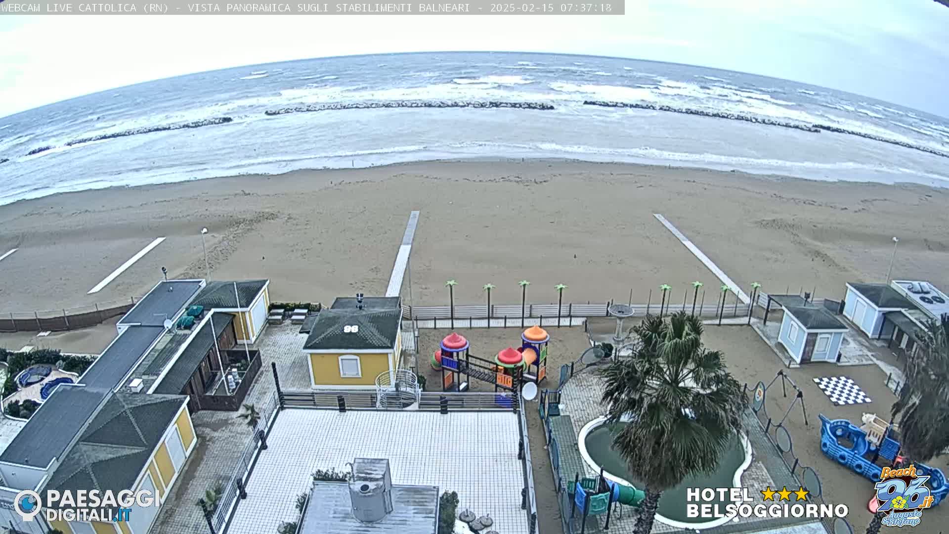 A beach with choppy waves under an overcast sky is viewed from above, showing beachside buildings, a playground, and a small pool.