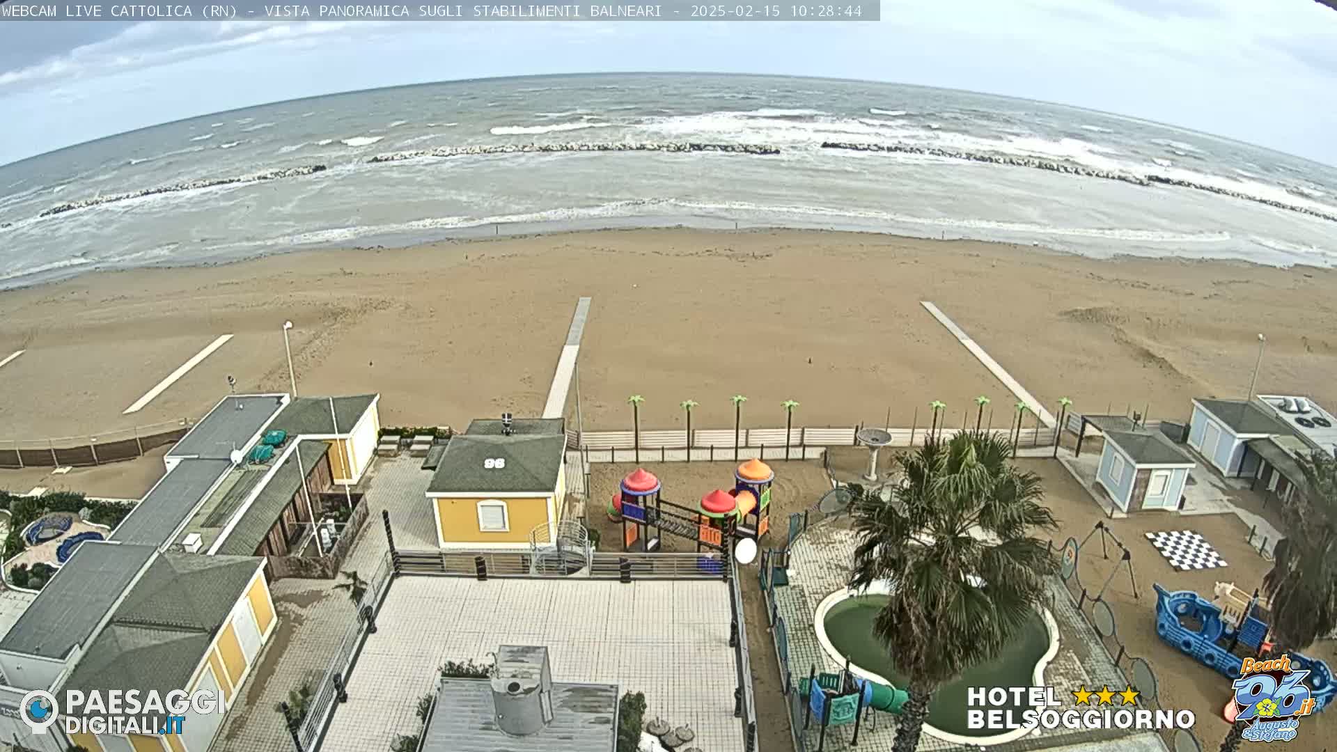 A beach with choppy waves under an overcast sky is viewed from above, showing beachside buildings, a playground, and a swimming pool.