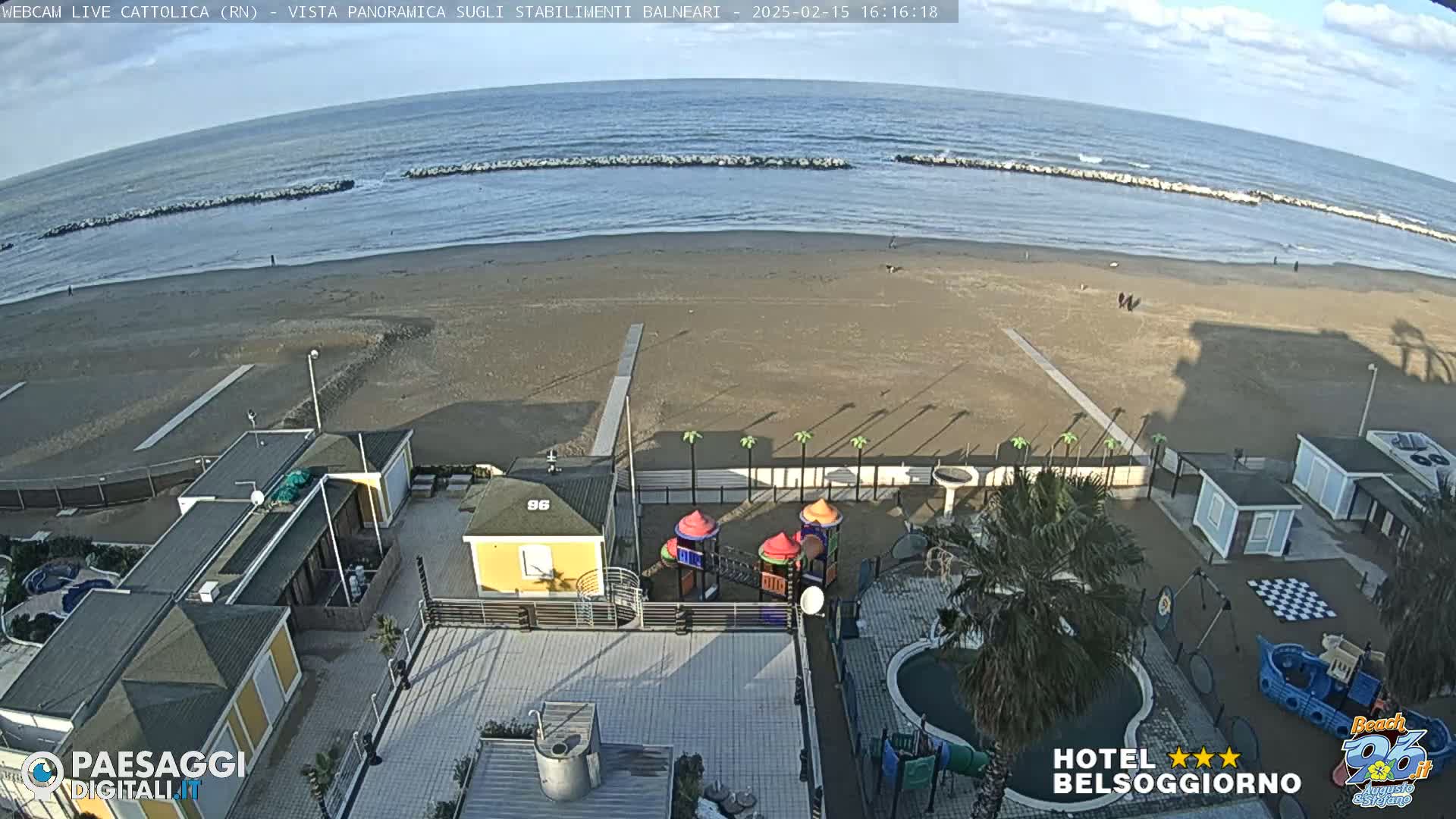 An aerial view shows a beach with a calm sea under a partly cloudy sky, along with beachside buildings, a playground, and a small swimming pool.