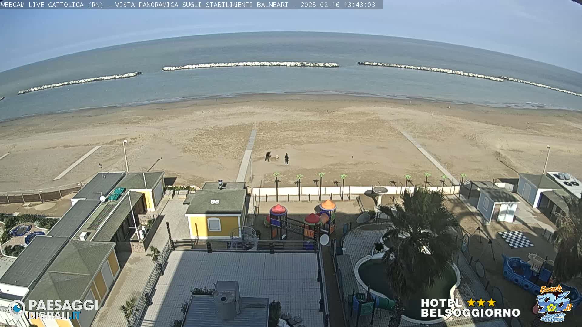 A sandy beach stretches to a calm sea, protected by breakwaters, with beachfront buildings, a playground, and a small pool visible in the foreground under a clear sky.