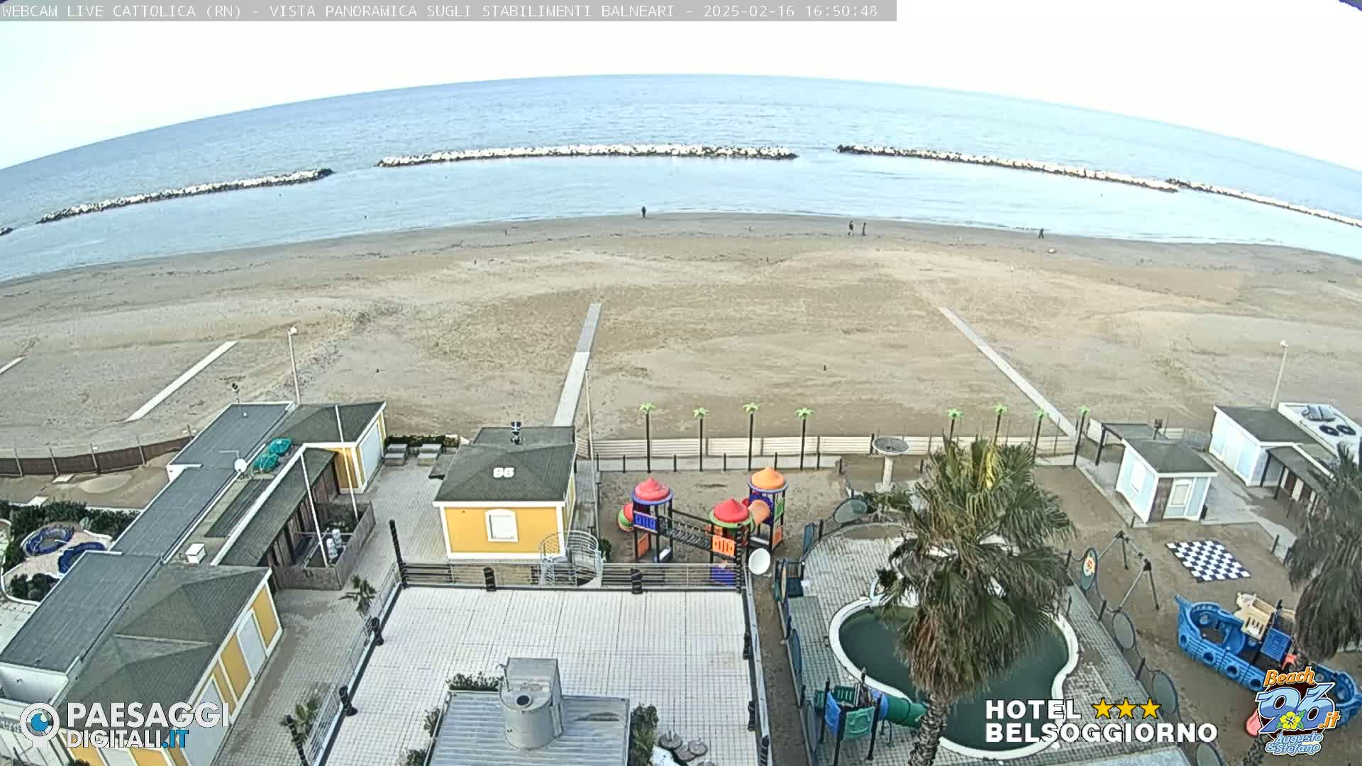 An aerial view of a beach with calm water, a sandy shoreline, beach buildings, a playground, and a swimming pool, under a cloudy sky.