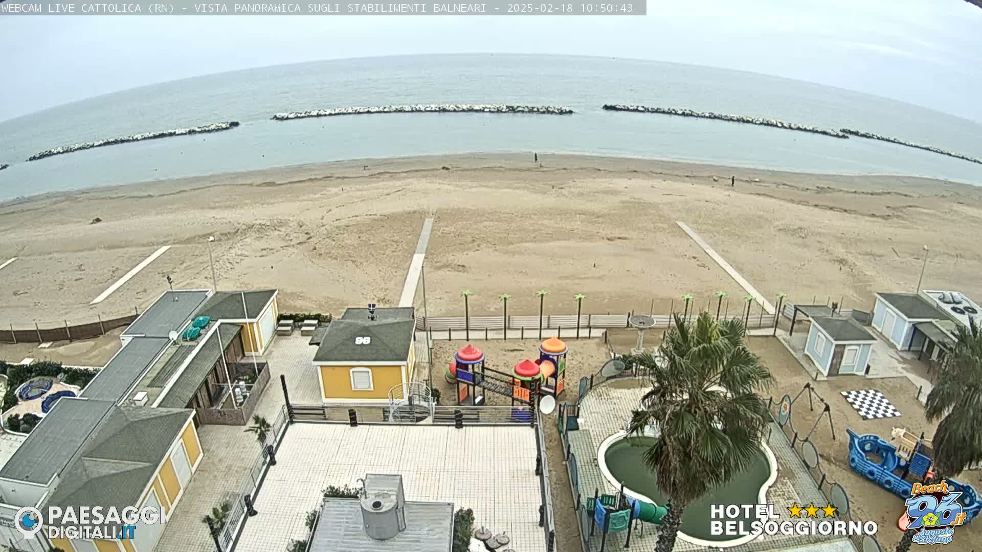 An overcast day shows a sandy beach with calm ocean water, breakwaters, beachside buildings, a playground, and a small pool.