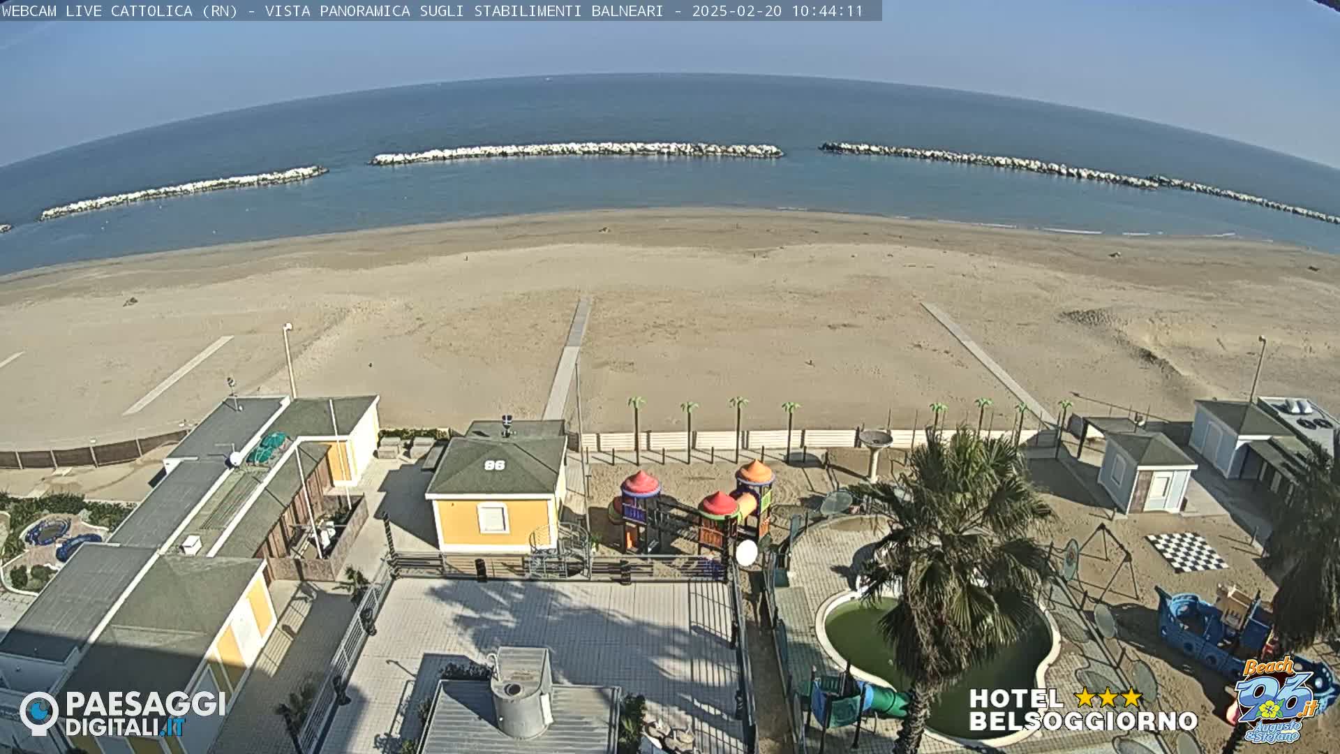 A sunny beach scene shows a sandy beach with calm ocean water, breakwaters, beachside buildings, a playground, and a small pool.