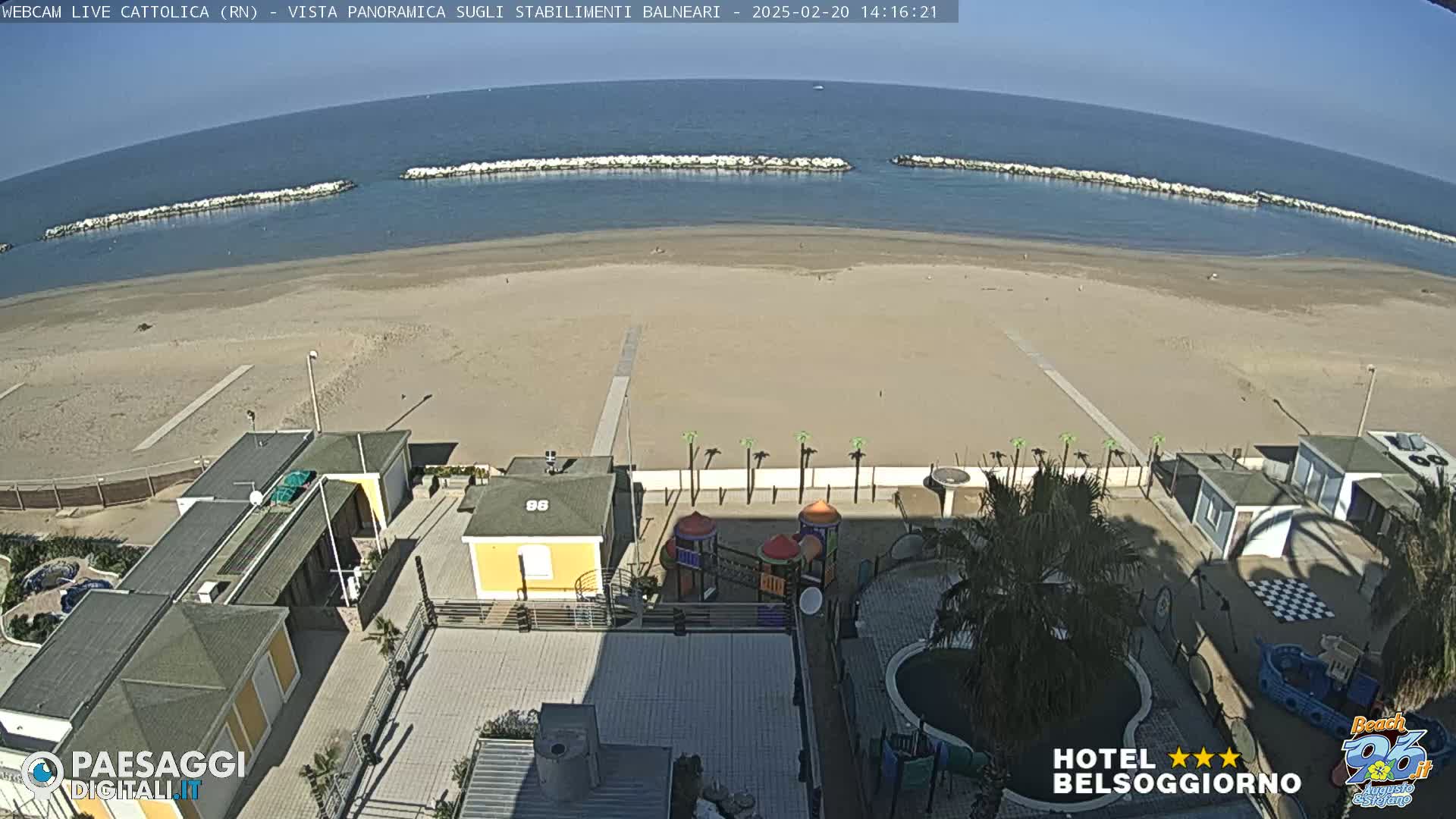 A mostly sunny day shows a sandy beach, calm ocean with breakwaters, and beachside buildings including a small playground and swimming pool.