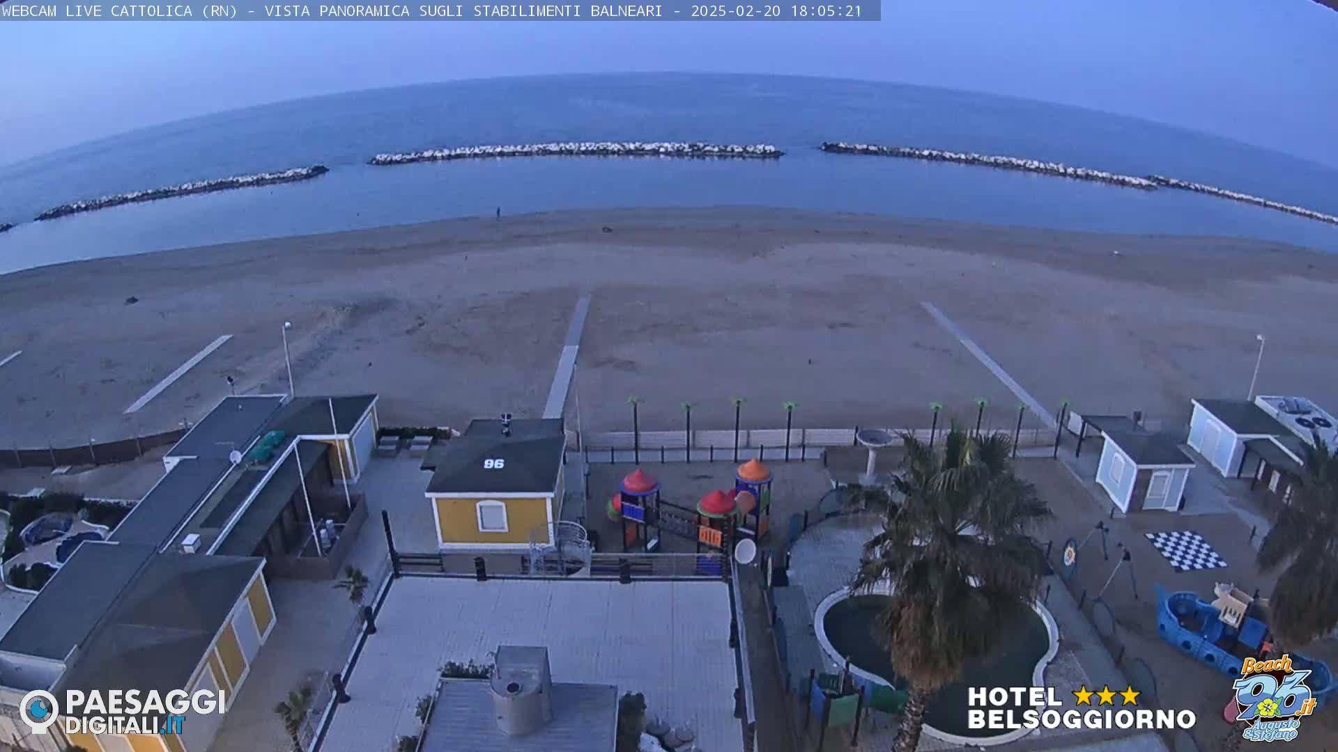 A beach with calm water and a few buildings, including a small playground, is seen from an elevated viewpoint at dusk.