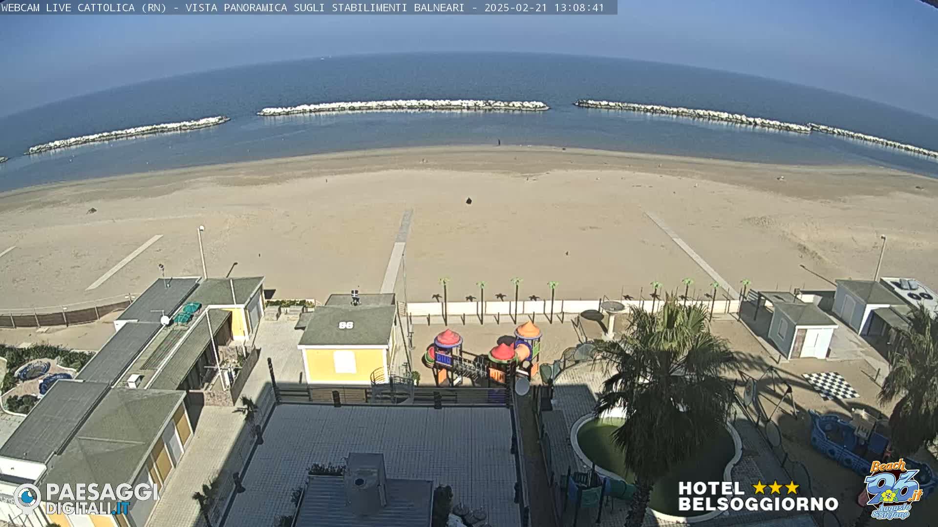 A beach with calm, clear water and a sandy shore, overlooked by buildings with a playground, under a sunny sky.