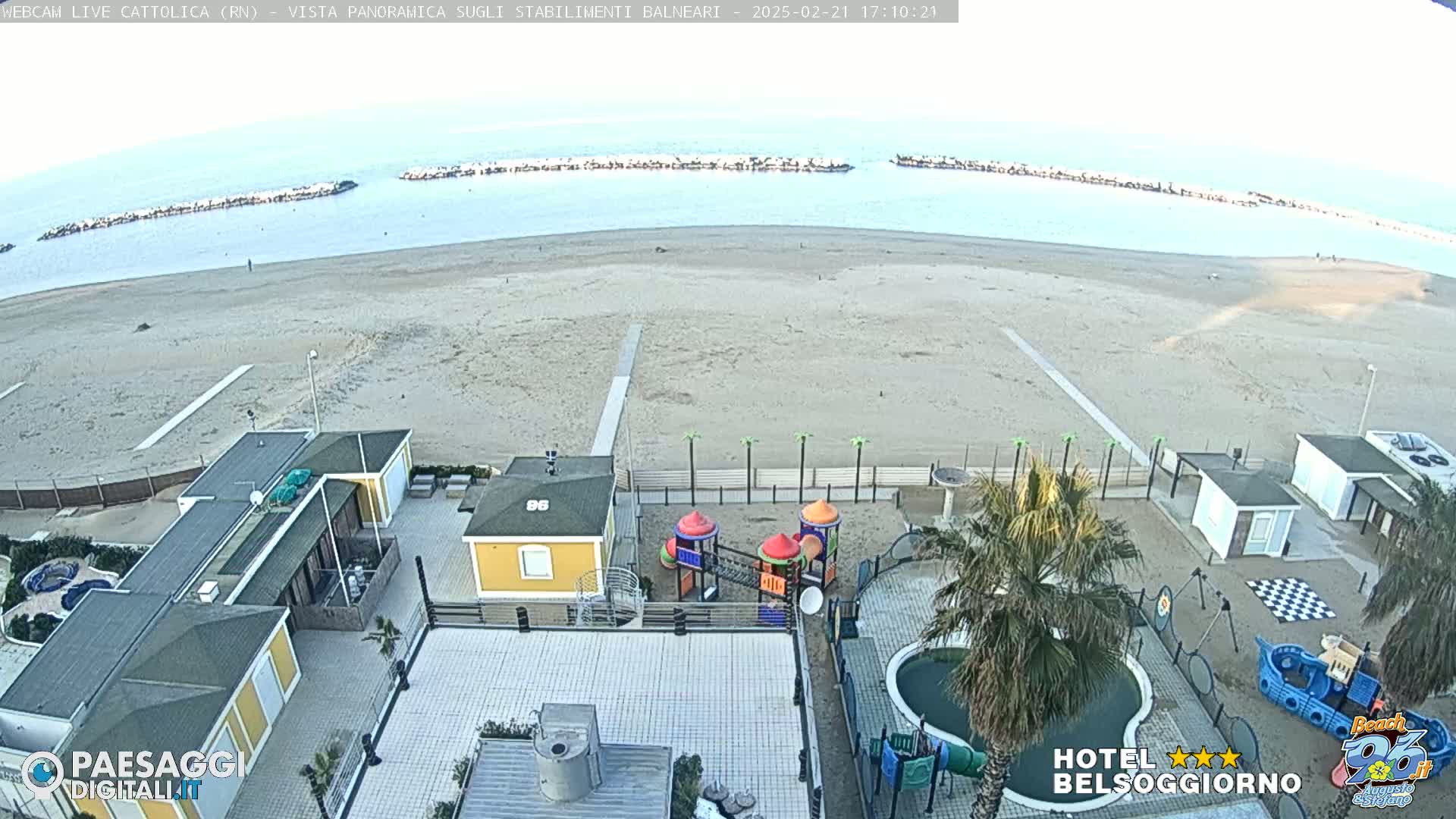 An overhead view of a sandy beach with calm water, breakwaters, beachside buildings, a playground, and a small pool, under a clear, bright sky.