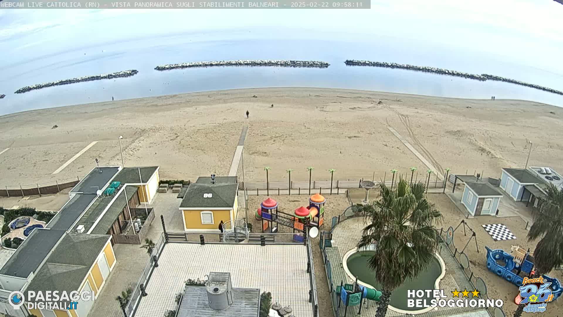 An aerial view of a sandy beach on an overcast day, showing beachside buildings, a playground, and a small pool.