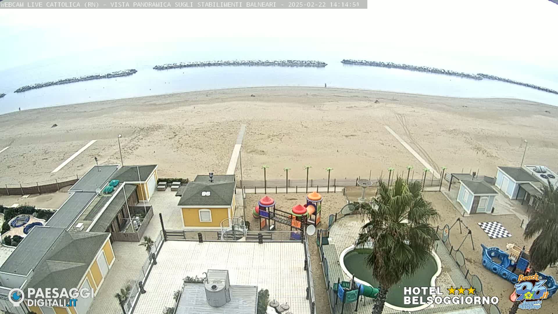 A sandy beach stretches out to a calm sea protected by breakwaters, under an overcast sky, with beachfront buildings and a playground visible in the foreground.