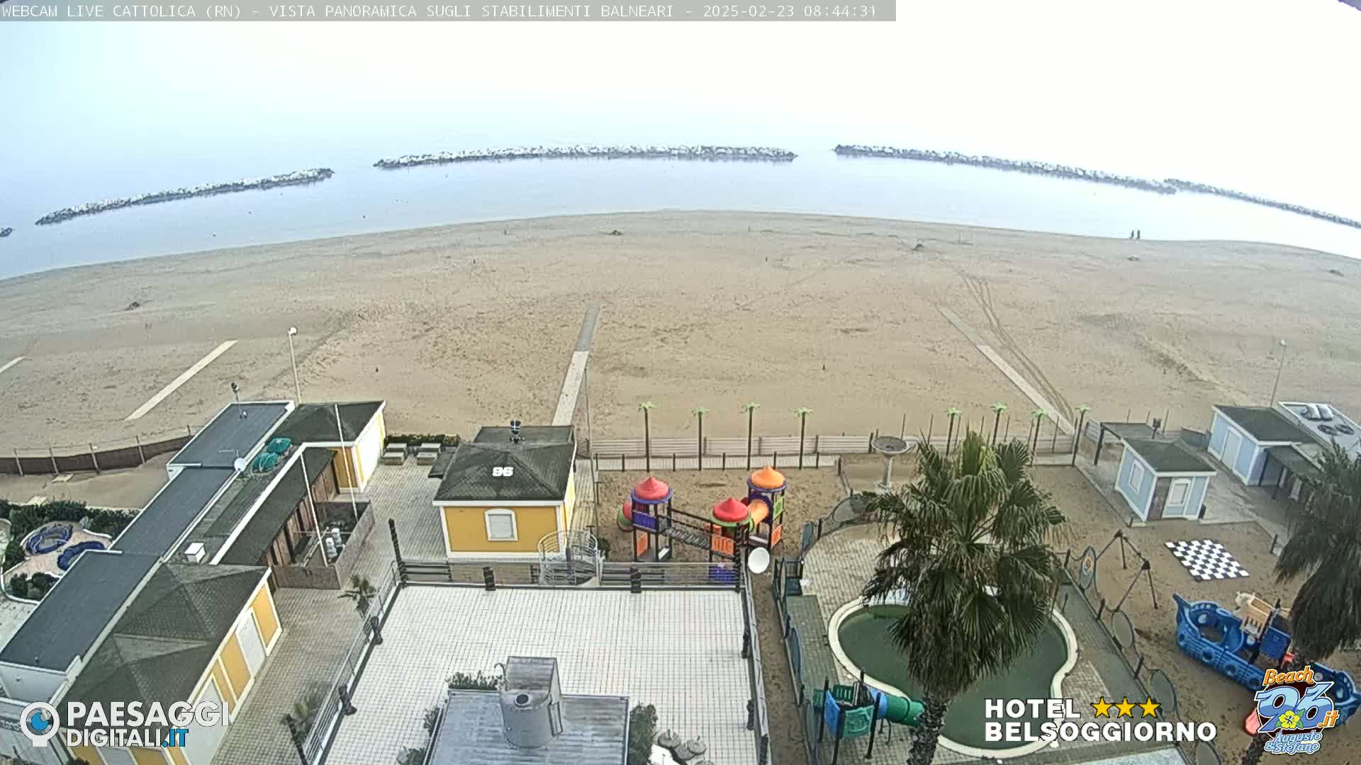 A sandy beach with calm, overcast water is seen from an elevated viewpoint, showing beachside buildings, a playground, and a small pool.