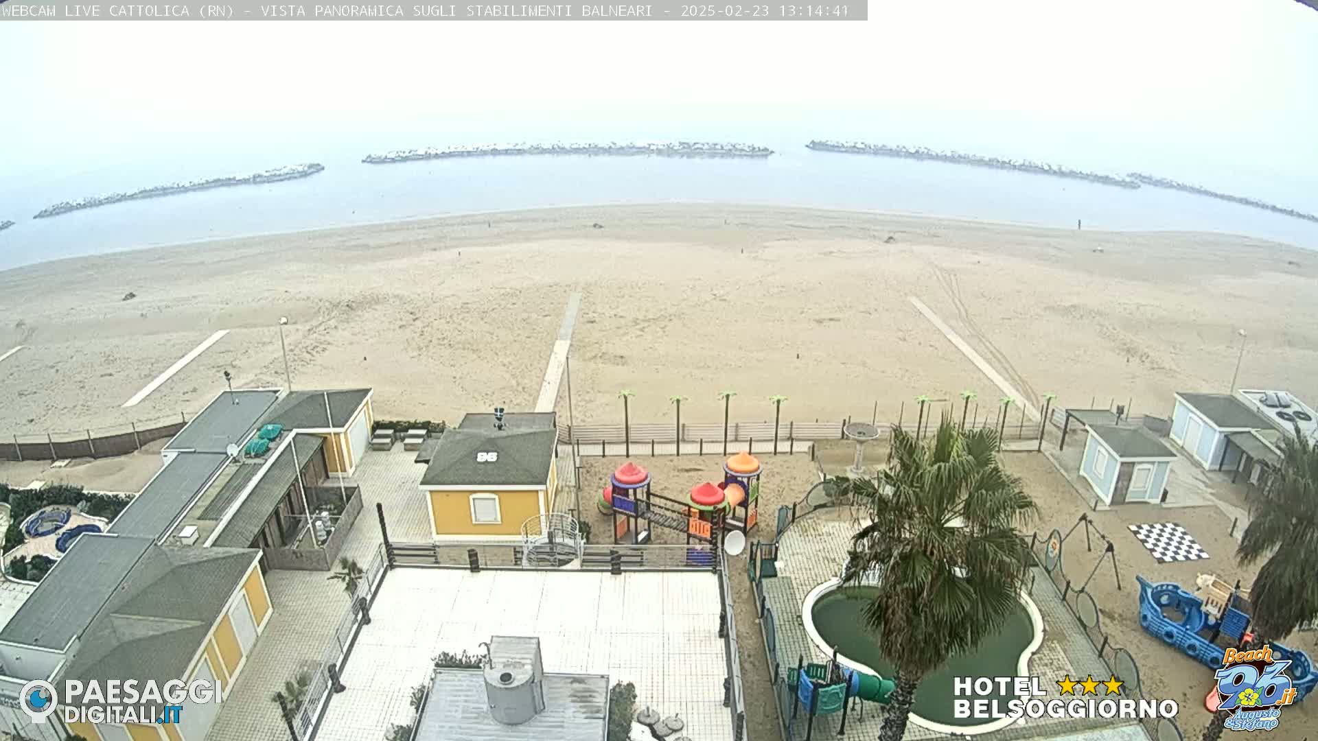 A mostly empty sandy beach stretches to a calm sea under an overcast sky, with beachside buildings, a playground, and a small pool visible in the foreground.