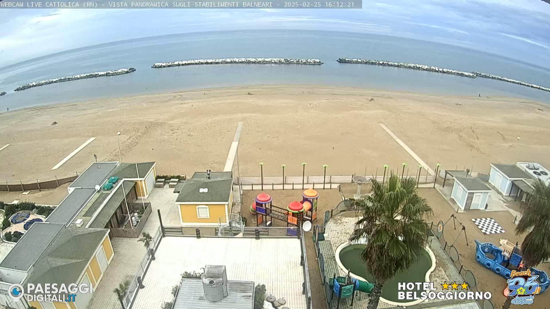 An aerial view of a sandy beach and beachfront buildings on an overcast day, showing a calm sea with breakwaters, a playground, and a small pool.