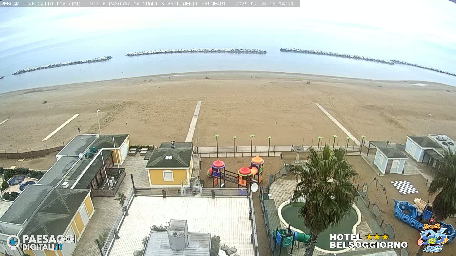An overcast day at a beach, viewed from above, shows a sandy beach, calm water, breakwaters, beachside buildings, a playground, and a small pool.