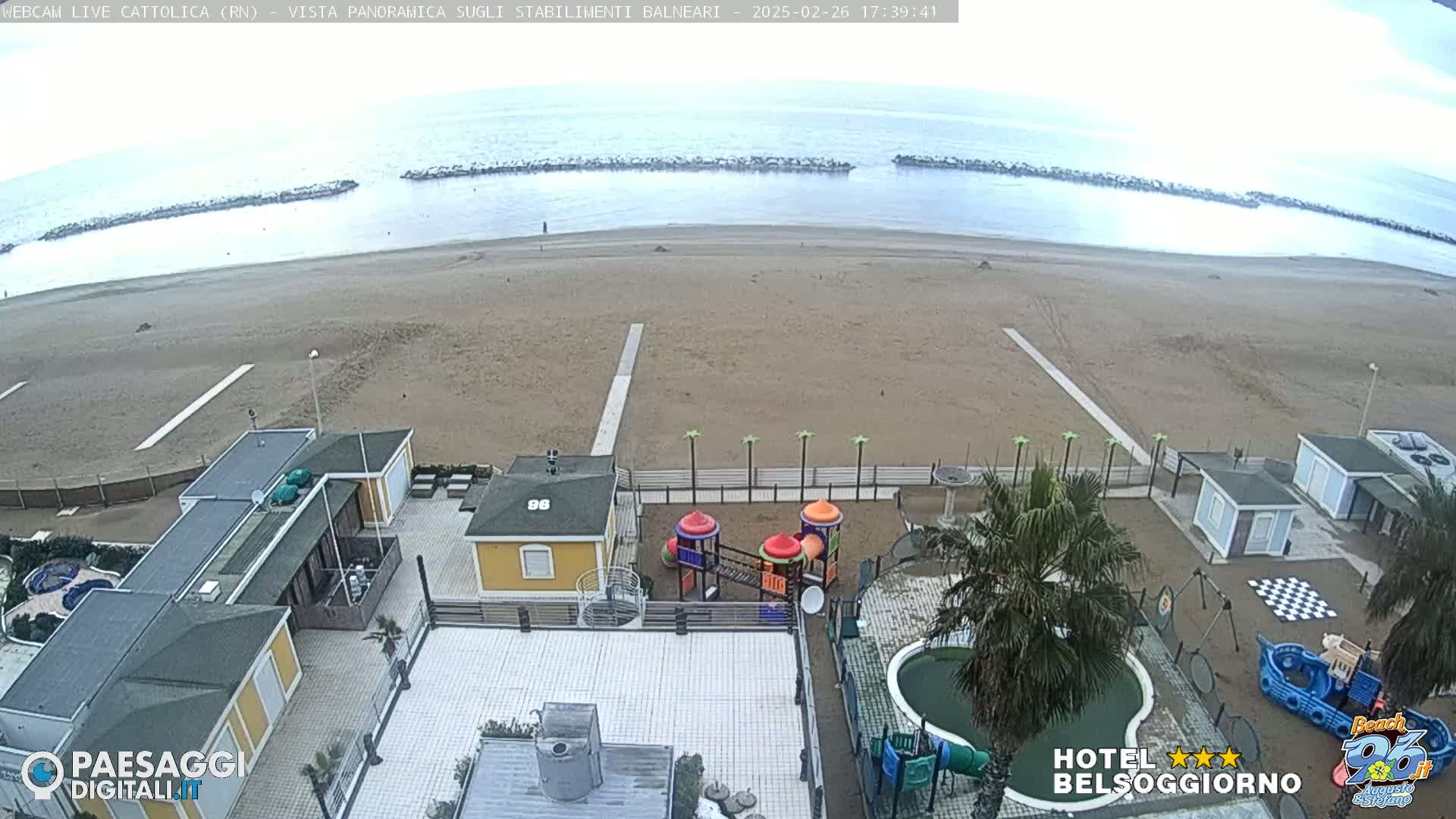 A beach scene on an overcast day shows a sandy beach with buildings, a playground, and a small pool, all situated near a calm ocean protected by breakwaters.