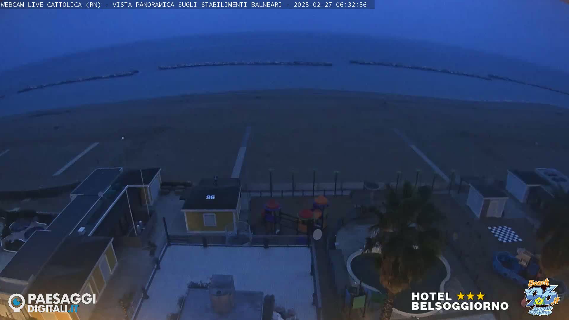 A high-angle, twilight view shows a beach with calm water and breakwaters in the distance, along with beachside buildings, a playground, and a small pool.