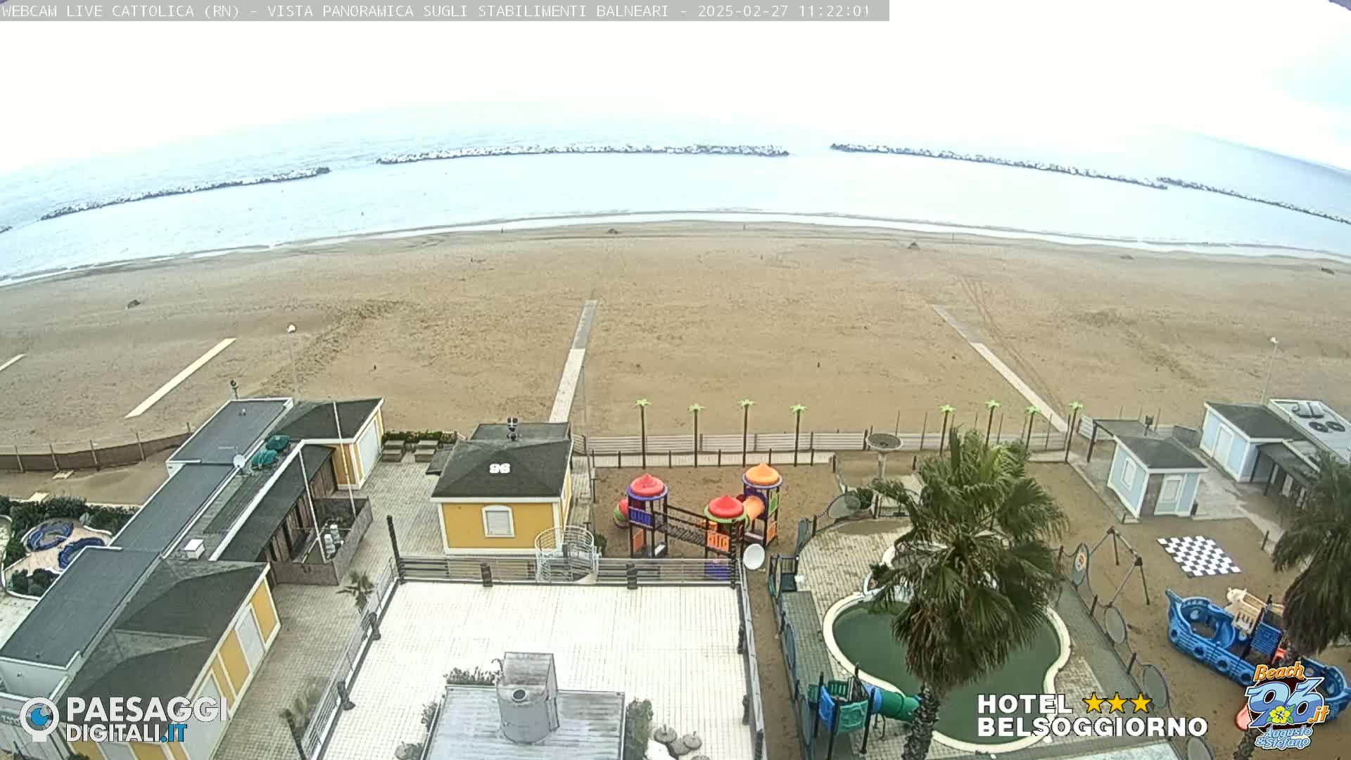 An overcast day at a beachside resort shows a sandy beach, calm ocean, and resort buildings with a playground and pool.