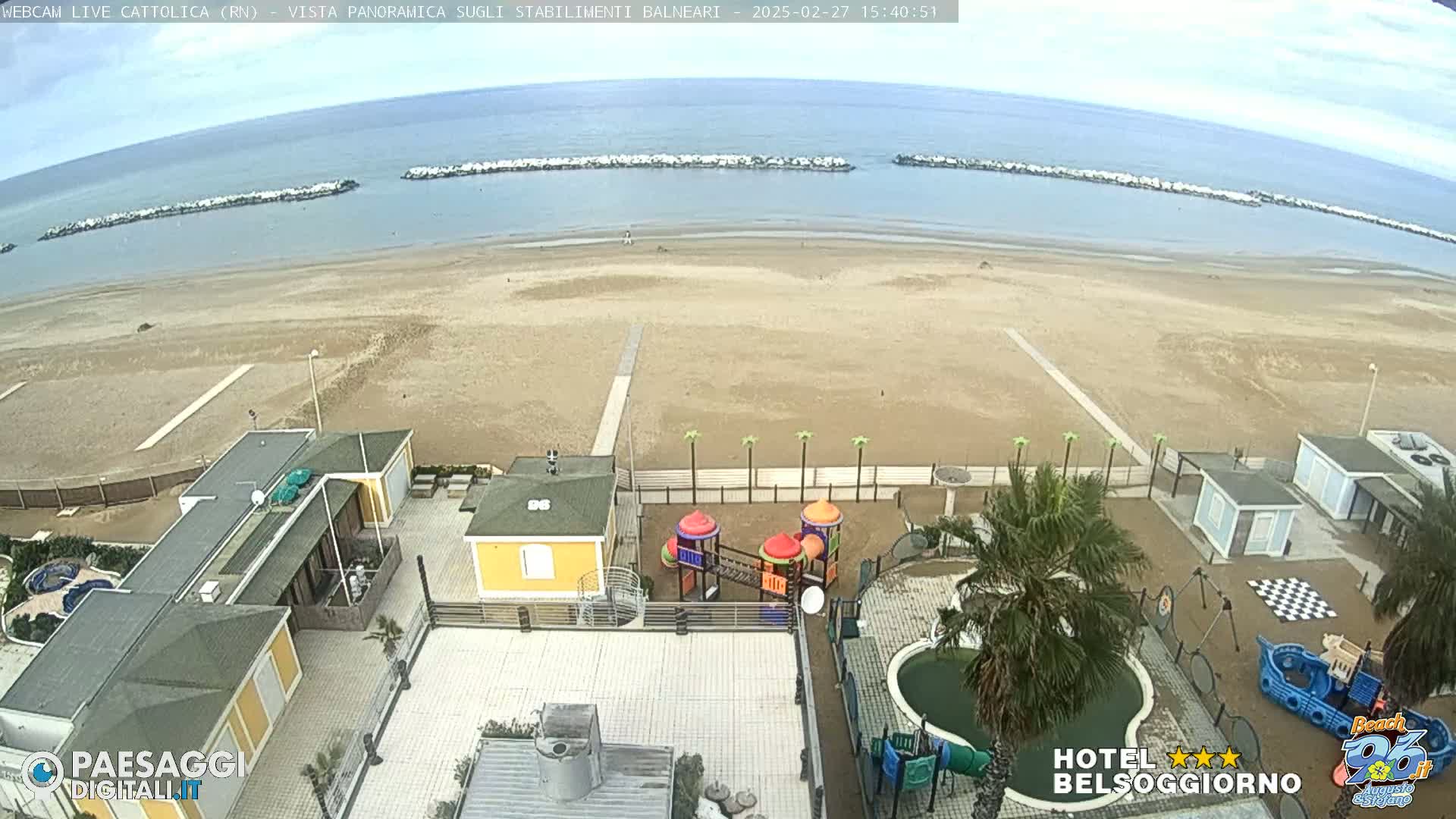 An expansive sandy beach with calm ocean water and breakwaters, viewed from above, showing beachside buildings, a playground, and a small pool under an overcast sky.