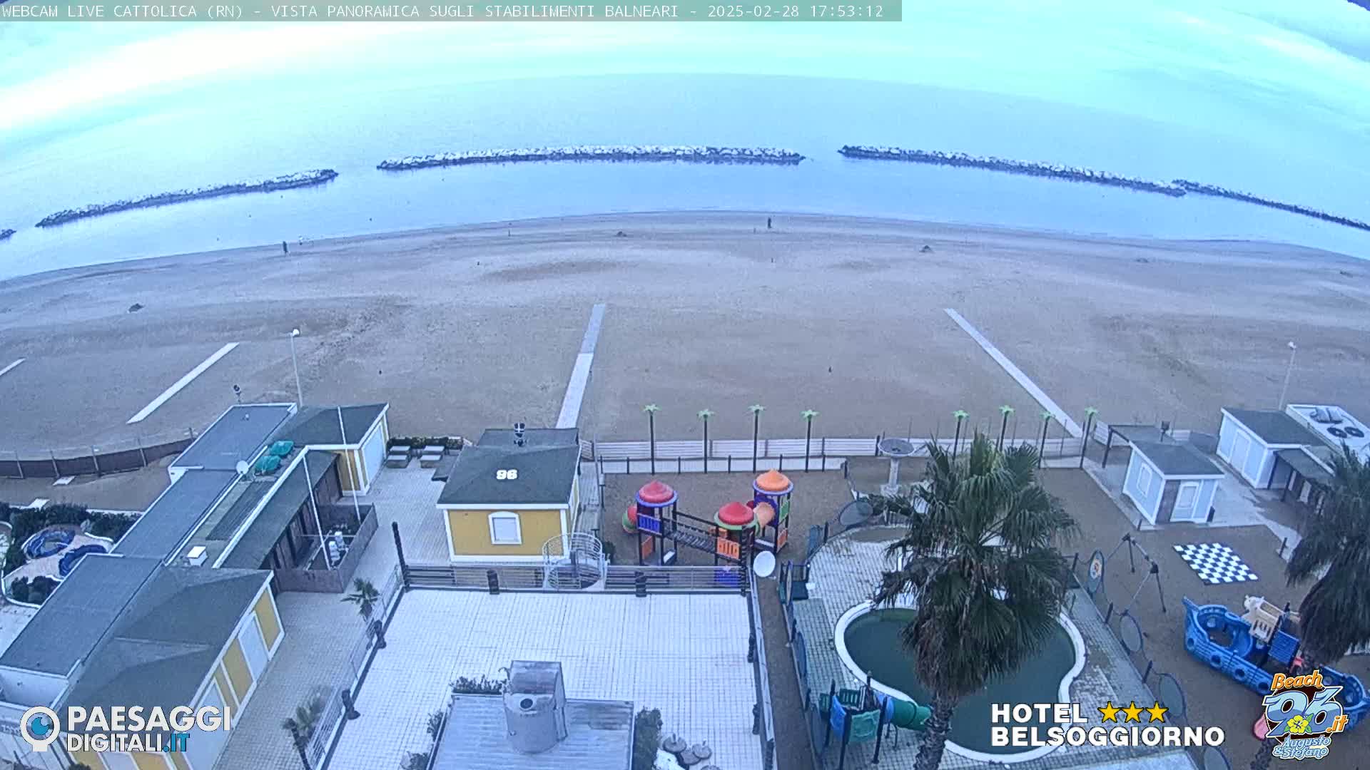 A beach with calm, light-blue water under a mostly clear, pale sky, viewed from above, showing beachside buildings, a playground, and a swimming pool.
