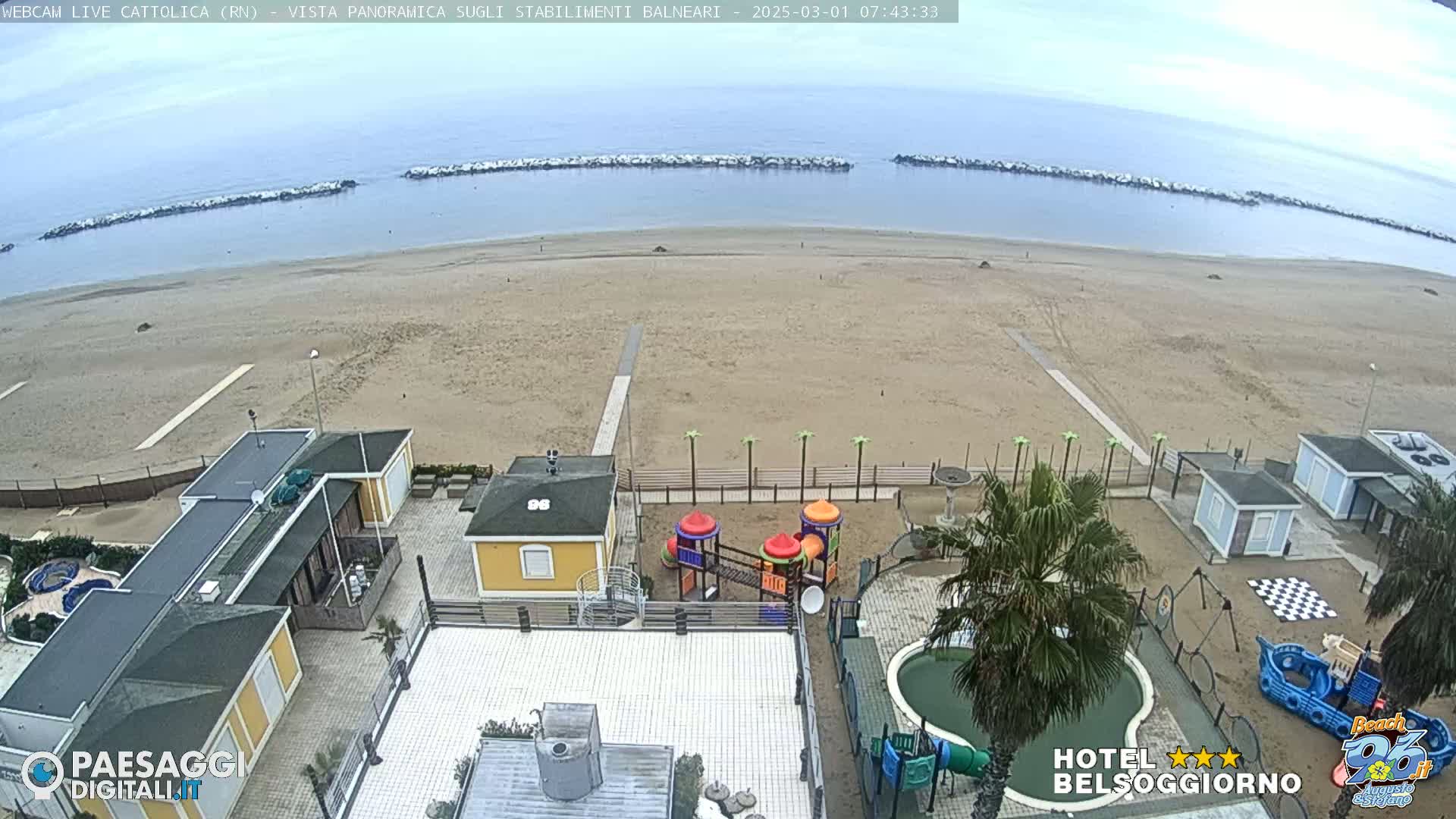 A sandy beach with calm ocean and breakwaters, viewed from above, showing beachfront buildings, a playground, and a small pool on an overcast day.