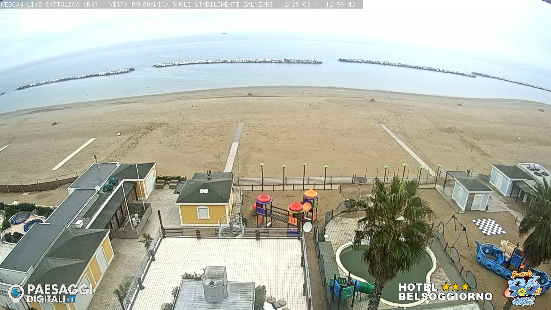 A sandy beach with calm, overcast water, beachside buildings, a playground, and a small pool is seen from an elevated vantage point.